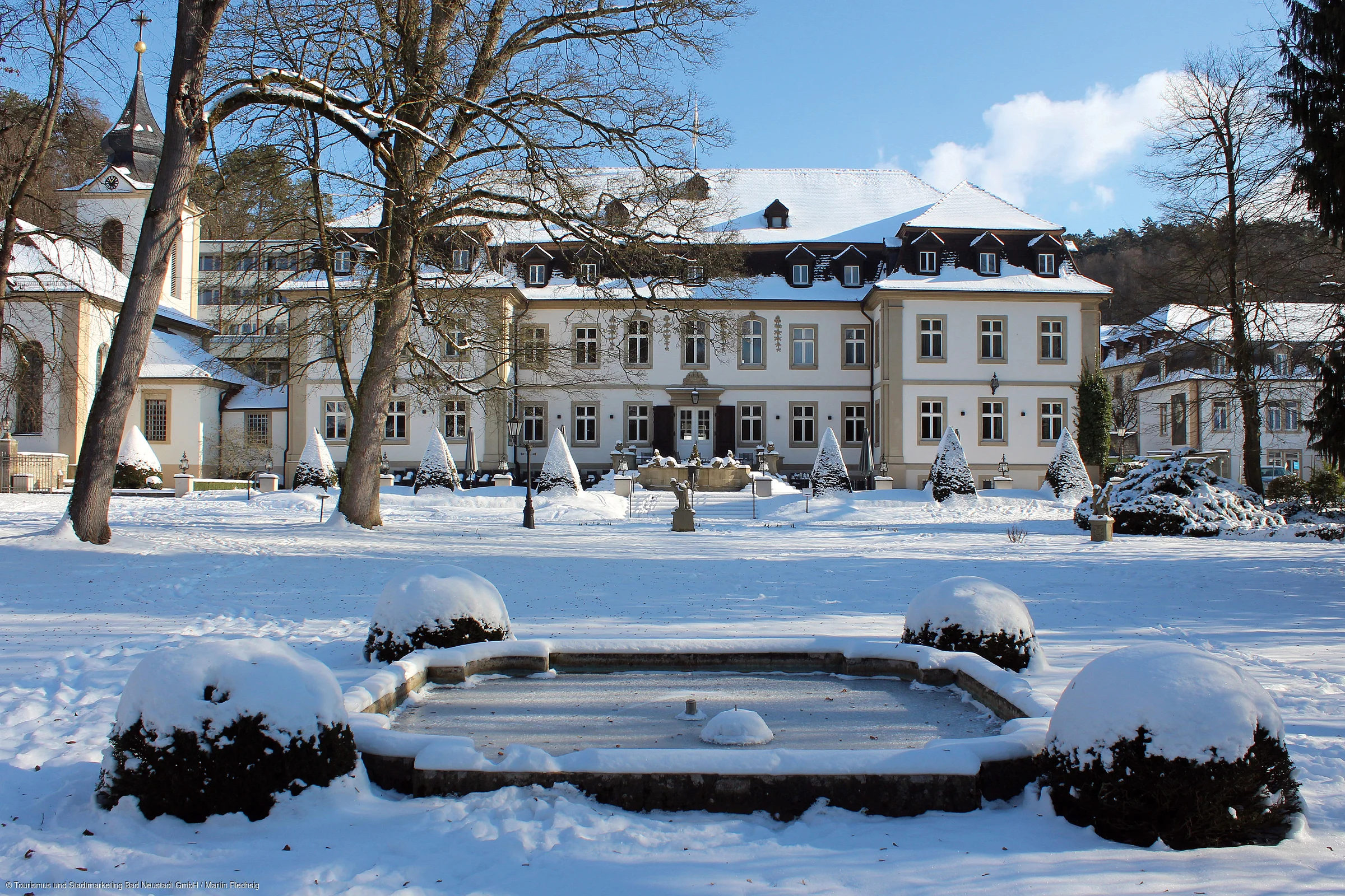Schneebedeckter Garten mit Brunnen vor einem großen historischen Gebäude und Kirche bei klarem Himmel.