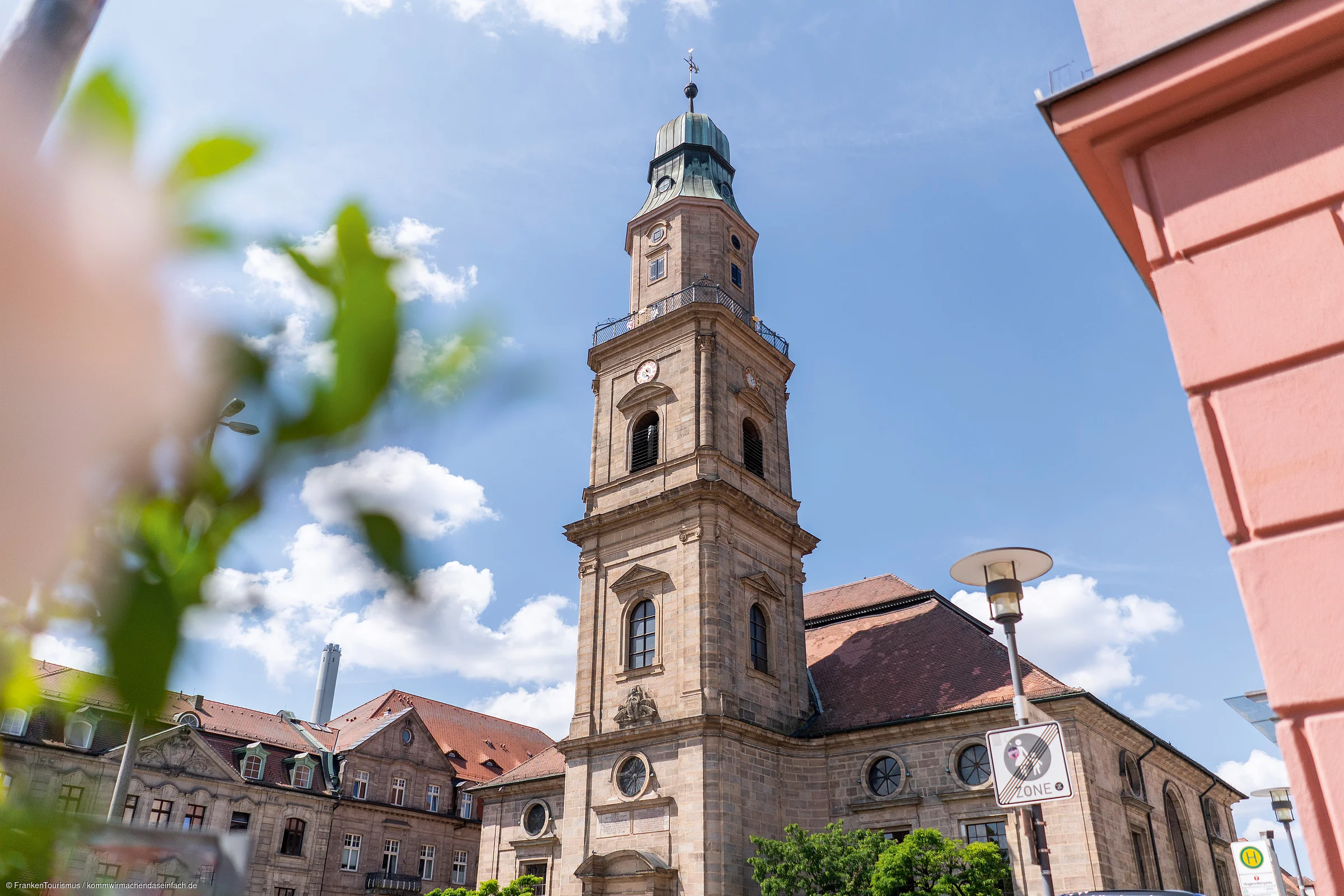Kirchturm eines historischen Gebäudes bei blauem Himmel, unscharfe Blätter im Vordergrund, Straßenschild sichtbar