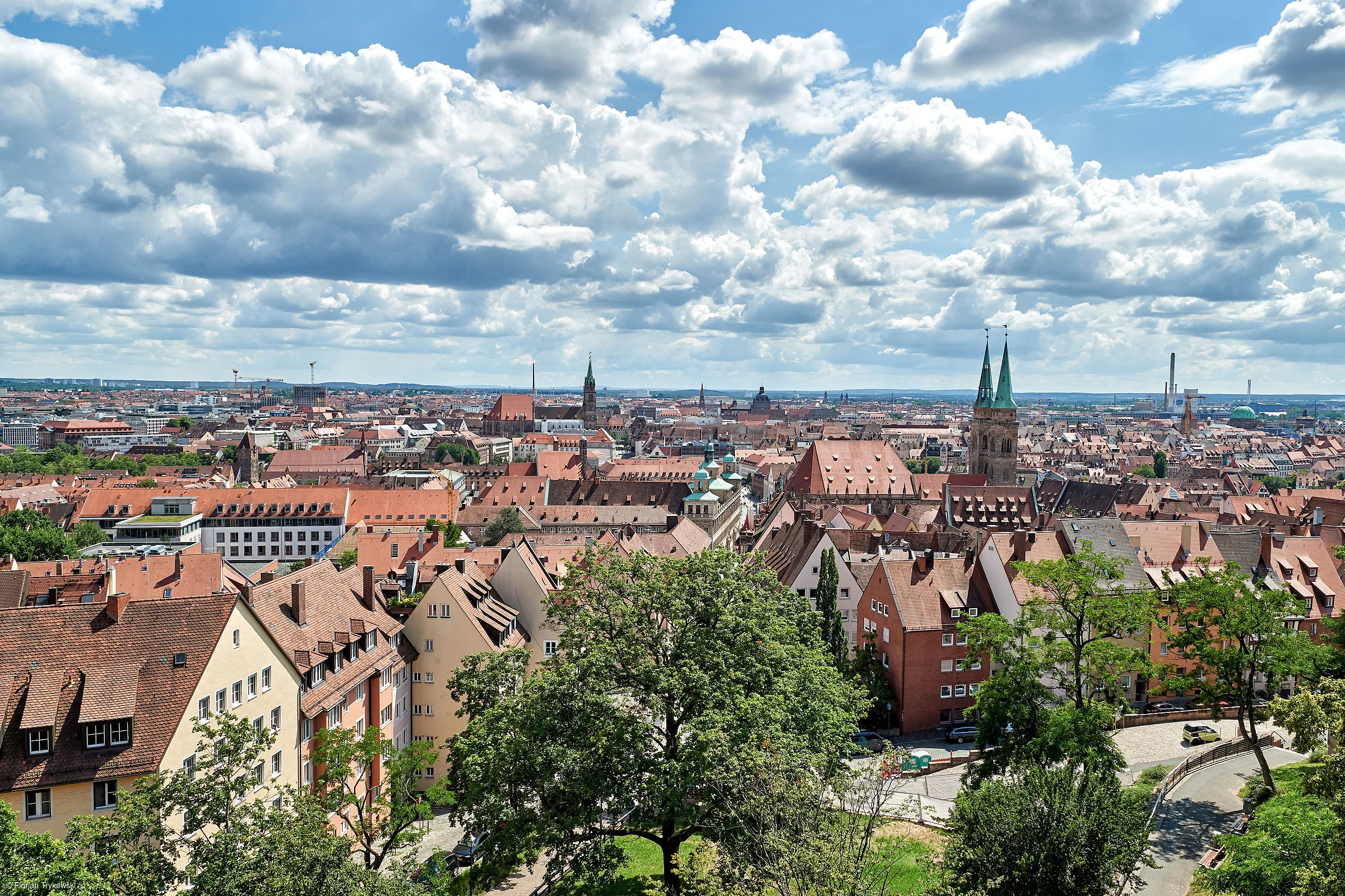 Blick über eine Stadt mit roten Dächern, Kirchen mit Türmen und bewölktem Himmel im Hintergrund.