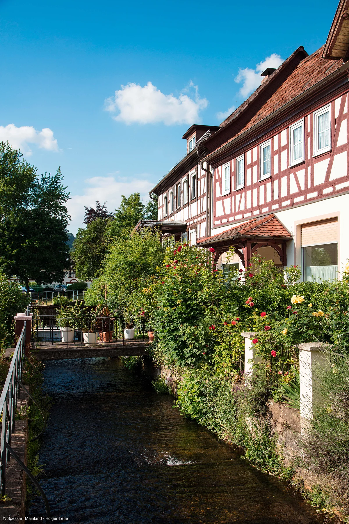 Fachwerkhaus mit Garten und Blumen neben einem Bach unter blauem Himmel mit wenigen Wolken.