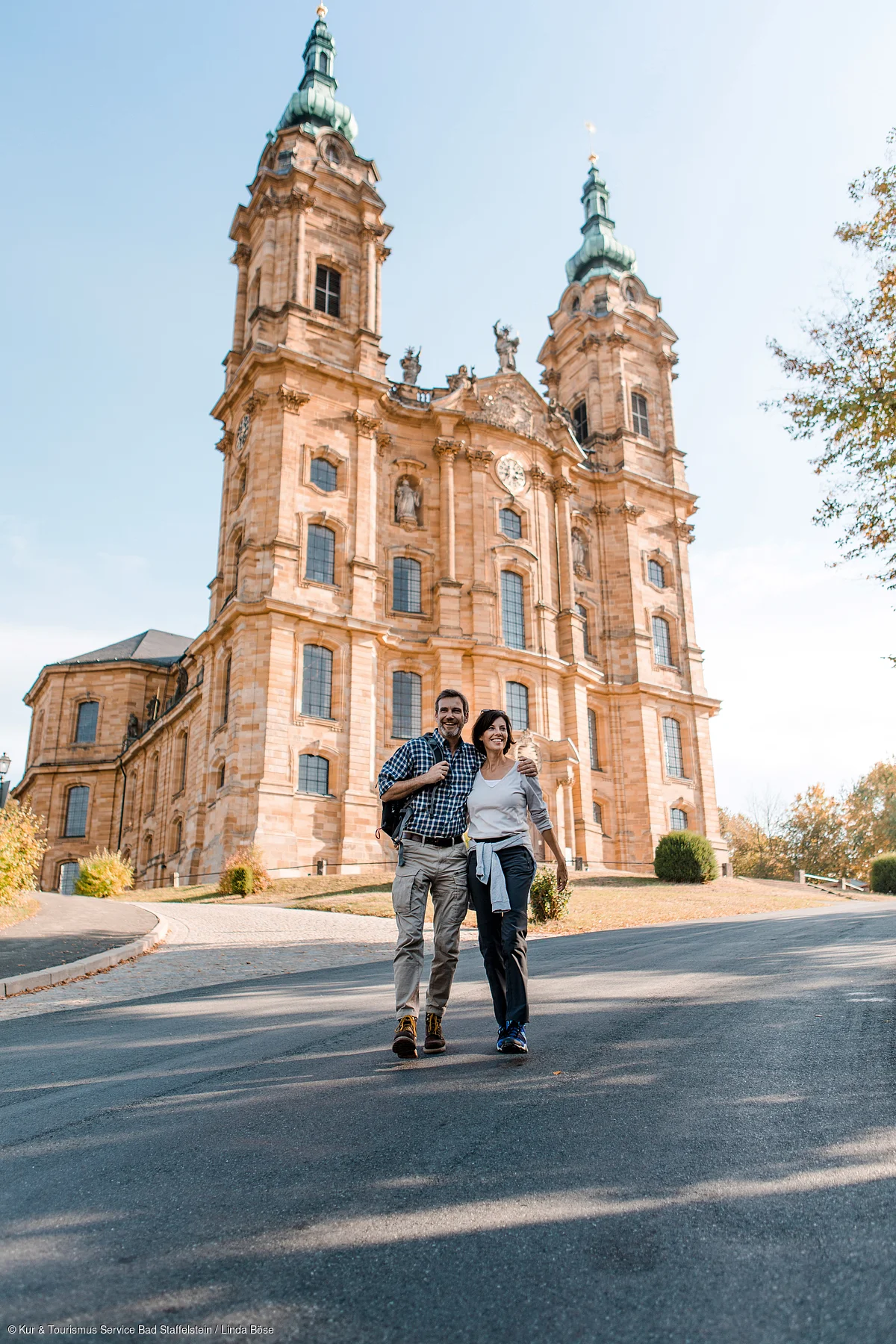 Paar mit Rucksack geht vor einer großen historischen Kirche mit zwei Türmen bei Tageslicht spazieren.
