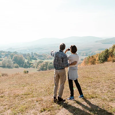 Zwei Personen stehen auf einer Wiese und blicken auf eine herbstliche Landschaft mit Hügeln und Bäumen.