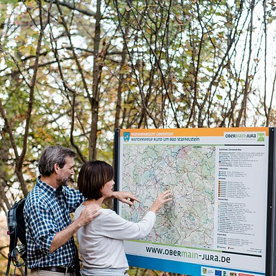 Zwei Wanderer lesen eine Wanderkarte an einem Infostand im Wald im Herbst.