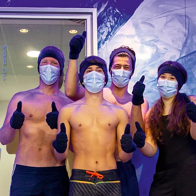 Vier Personen mit Mundschutz, Mütze und Handschuhen zeigen Daumen hoch vor einem Eisraum mit der Aufschrift „icelab -110° Celsius“.