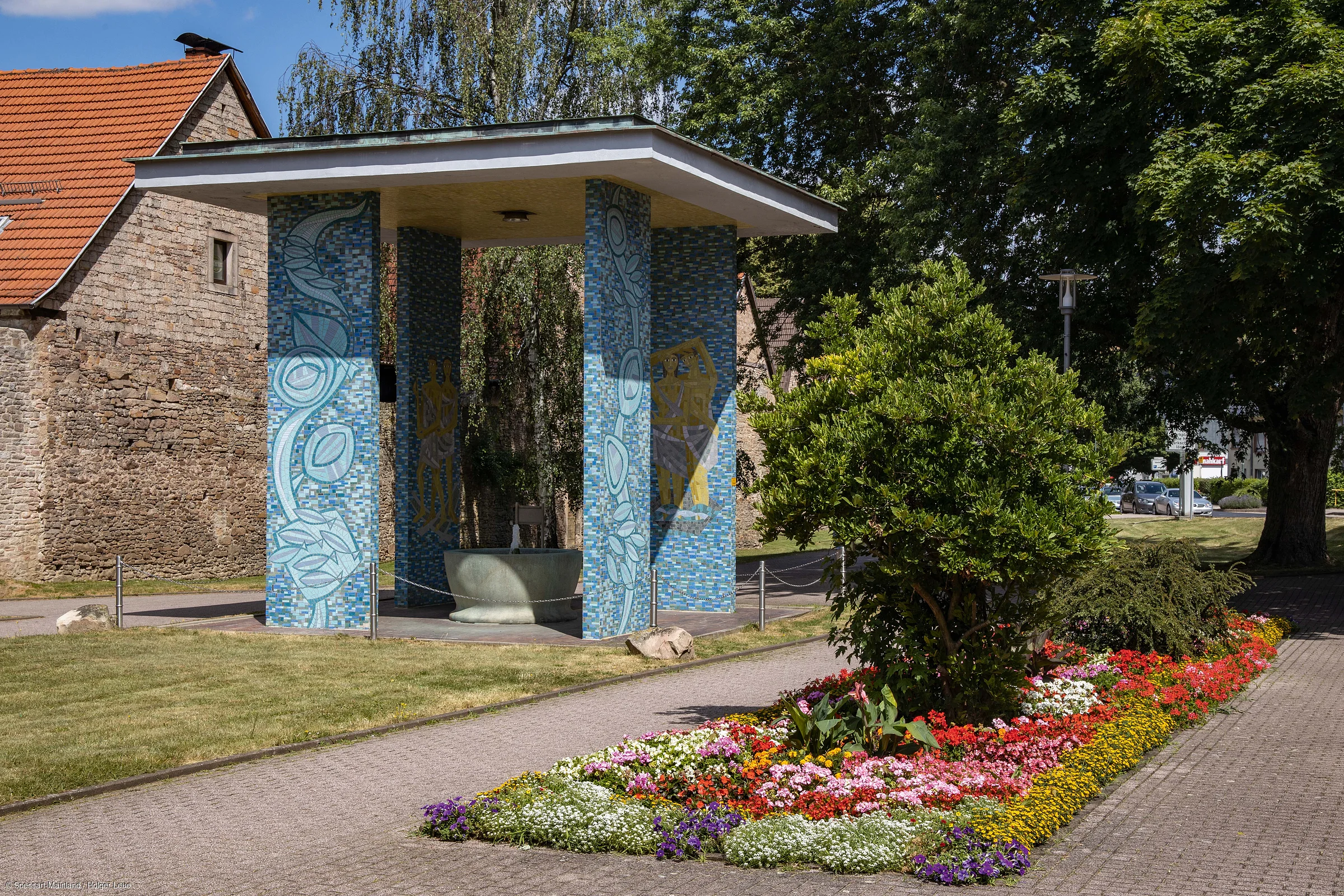 Pavillon mit blauen Mosaik-Säulen und Brunnen neben einem Blumenbeet und einem gepflasterten Weg bei Sonnenschein