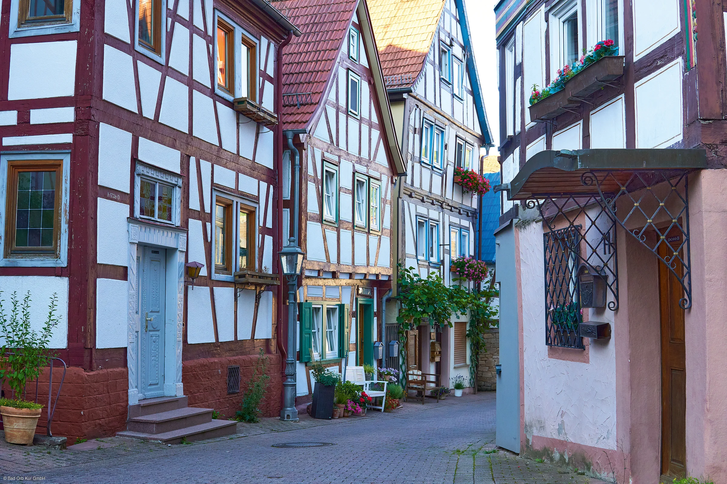 Enge Gasse mit Fachwerkhäusern, Blumentöpfen und Bänken an den Hauswänden bei Tageslicht.