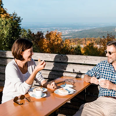 Zwei Personen sitzen draußen an einem Holztisch, essen Kuchen und trinken Kaffee, im Hintergrund Herbstwald.