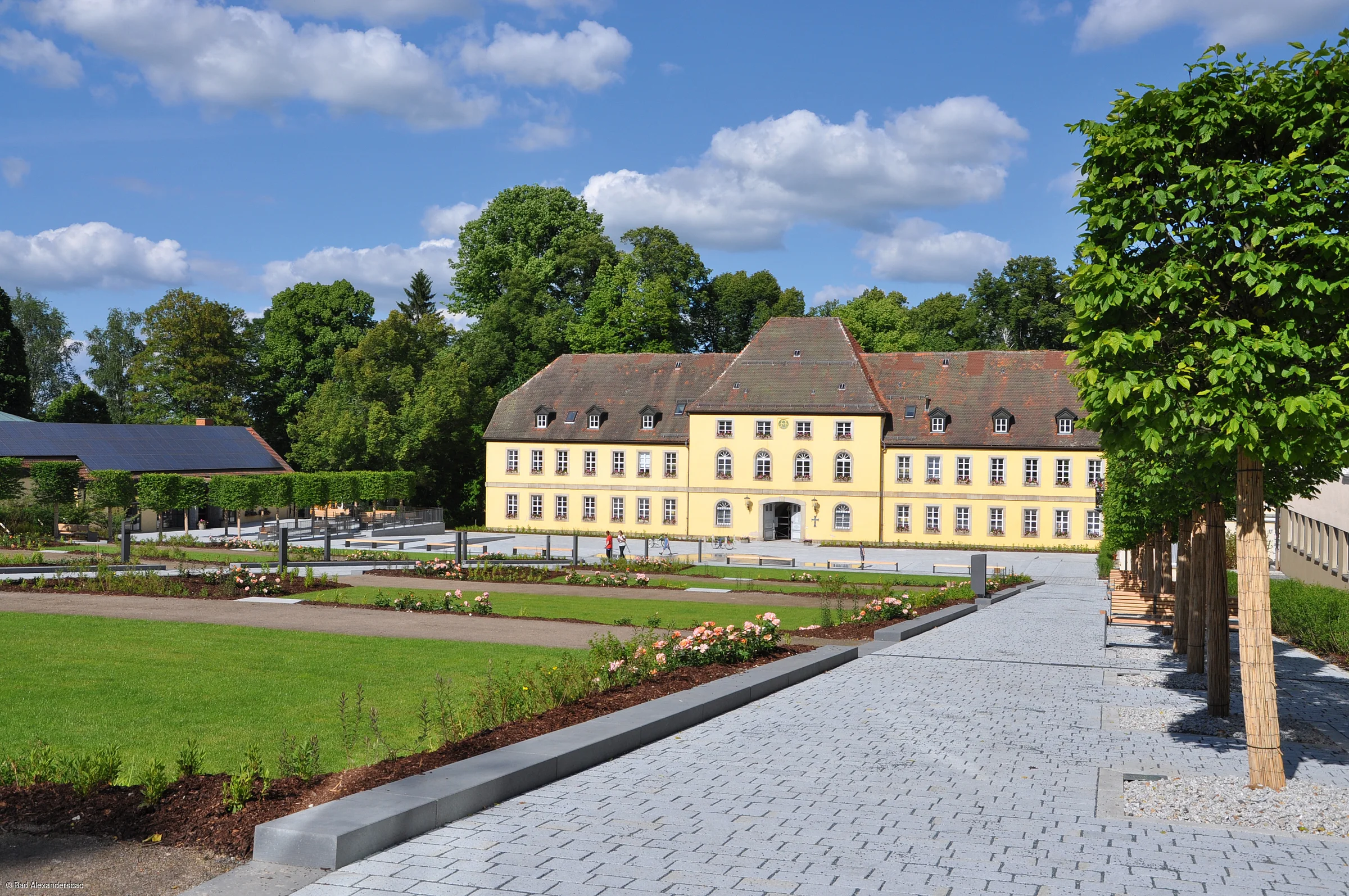 Gelbes historisches Gebäude mit großem Garten, gepflastertem Weg und Bäumen unter blauem Himmel mit Wolken.