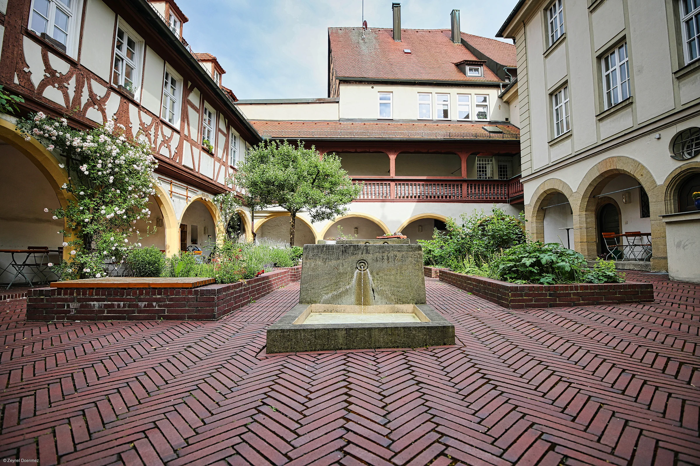Innenhof mit Backsteinboden, Brunnen in der Mitte, Blumenbeeten und Fachwerkhäusern mit Arkaden.