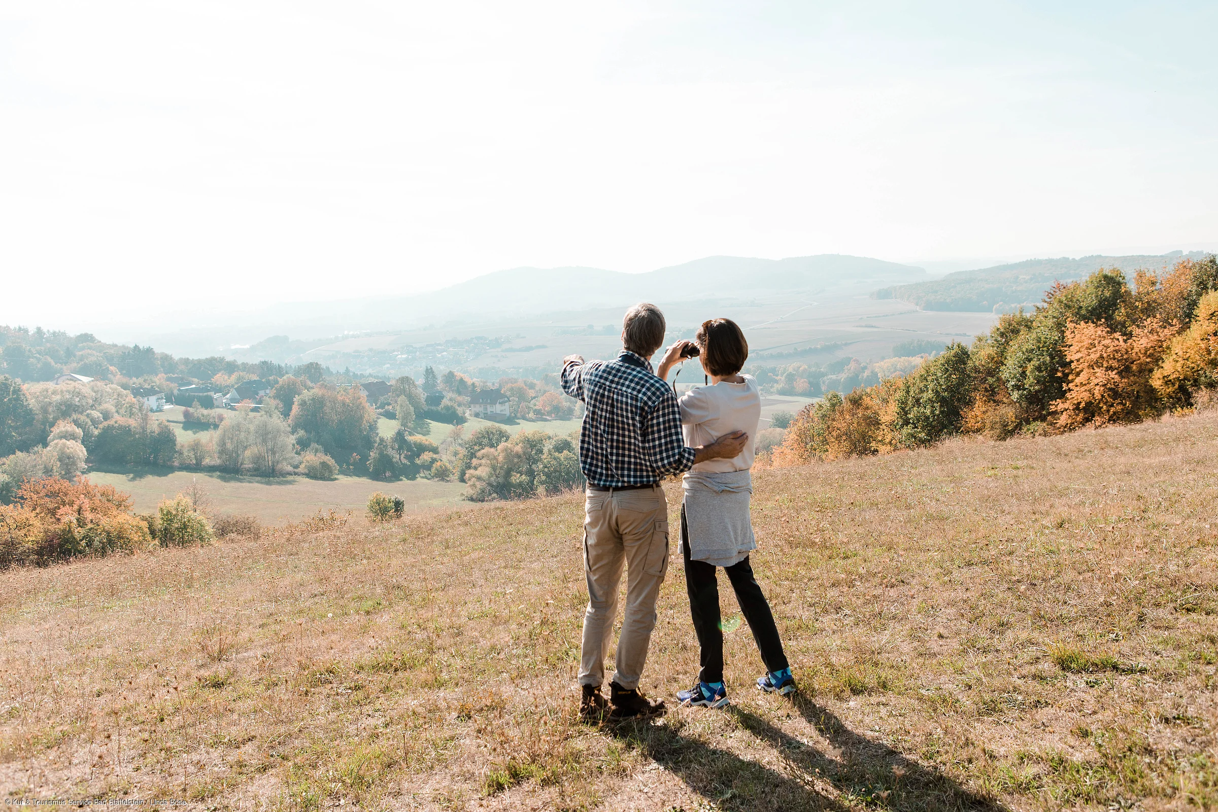 Zwei Personen stehen auf einer Wiese und blicken auf eine herbstliche Landschaft mit Hügeln und Bäumen.