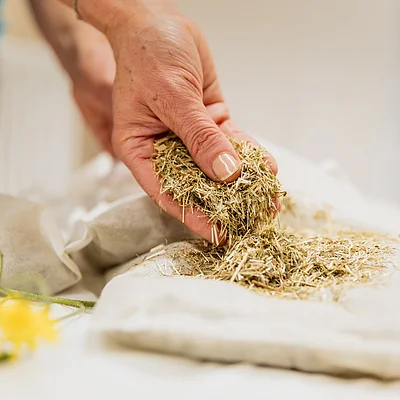 Hand hält getrocknete Kräuter über einem Stoffbeutel, gelbe Blume liegt daneben auf weißer Oberfläche.