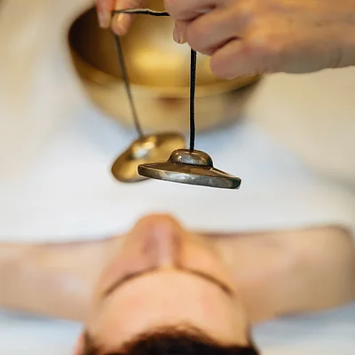 Person liegt mit geschlossenen Augen, darüber werden kleine Klangschalen an Schnüren gehalten.