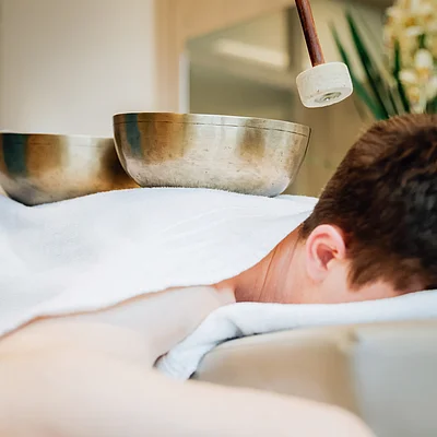 Person liegt mit Kopf nach unten auf Massageliege, Klangschalen und Schlägel auf Rücken, Pflanzen im Hintergrund