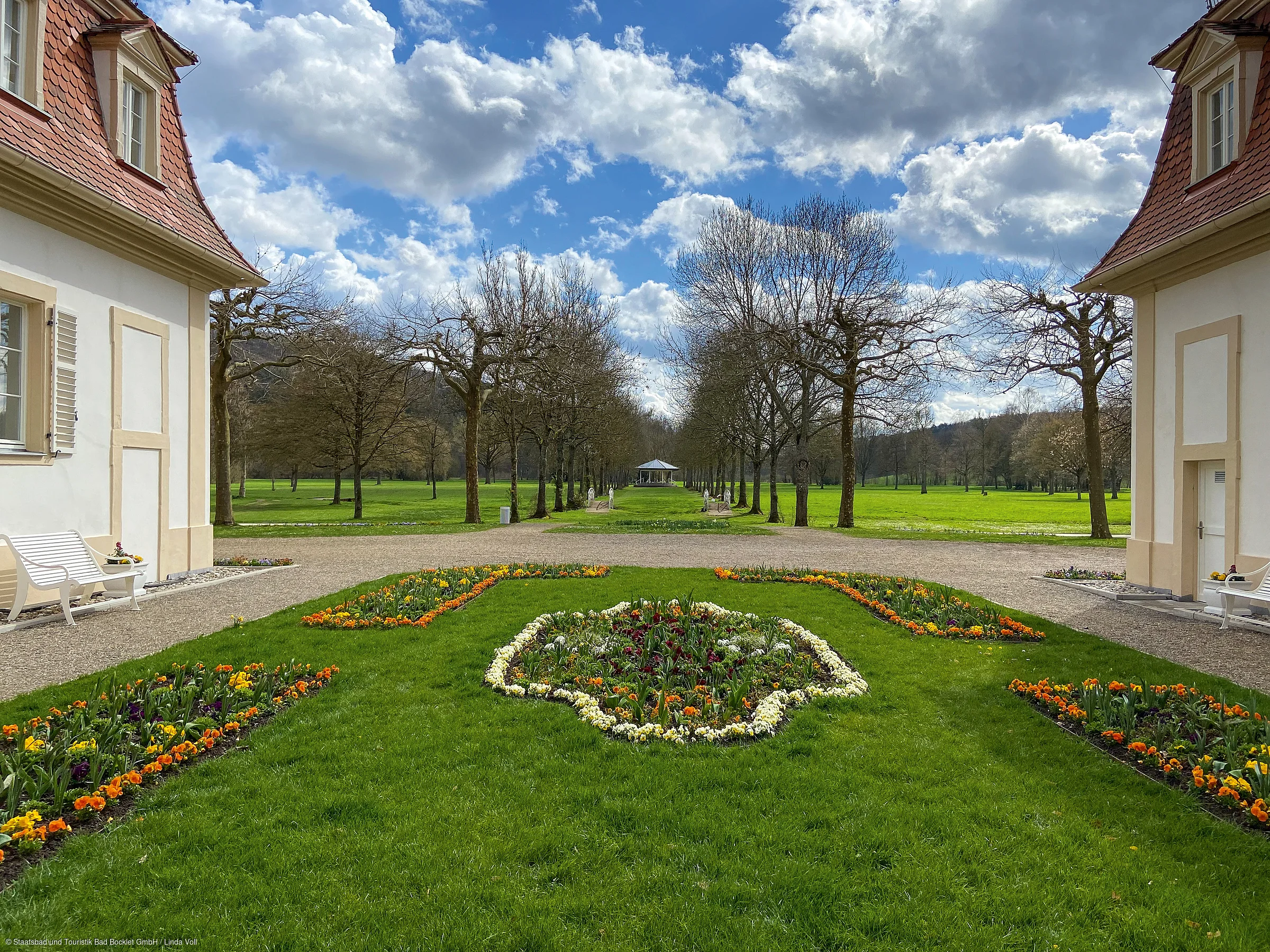 Blick auf symmetrischen Garten mit Blumenbeeten, Bänken, kahlen Bäumen und blauem Himmel mit Wolken