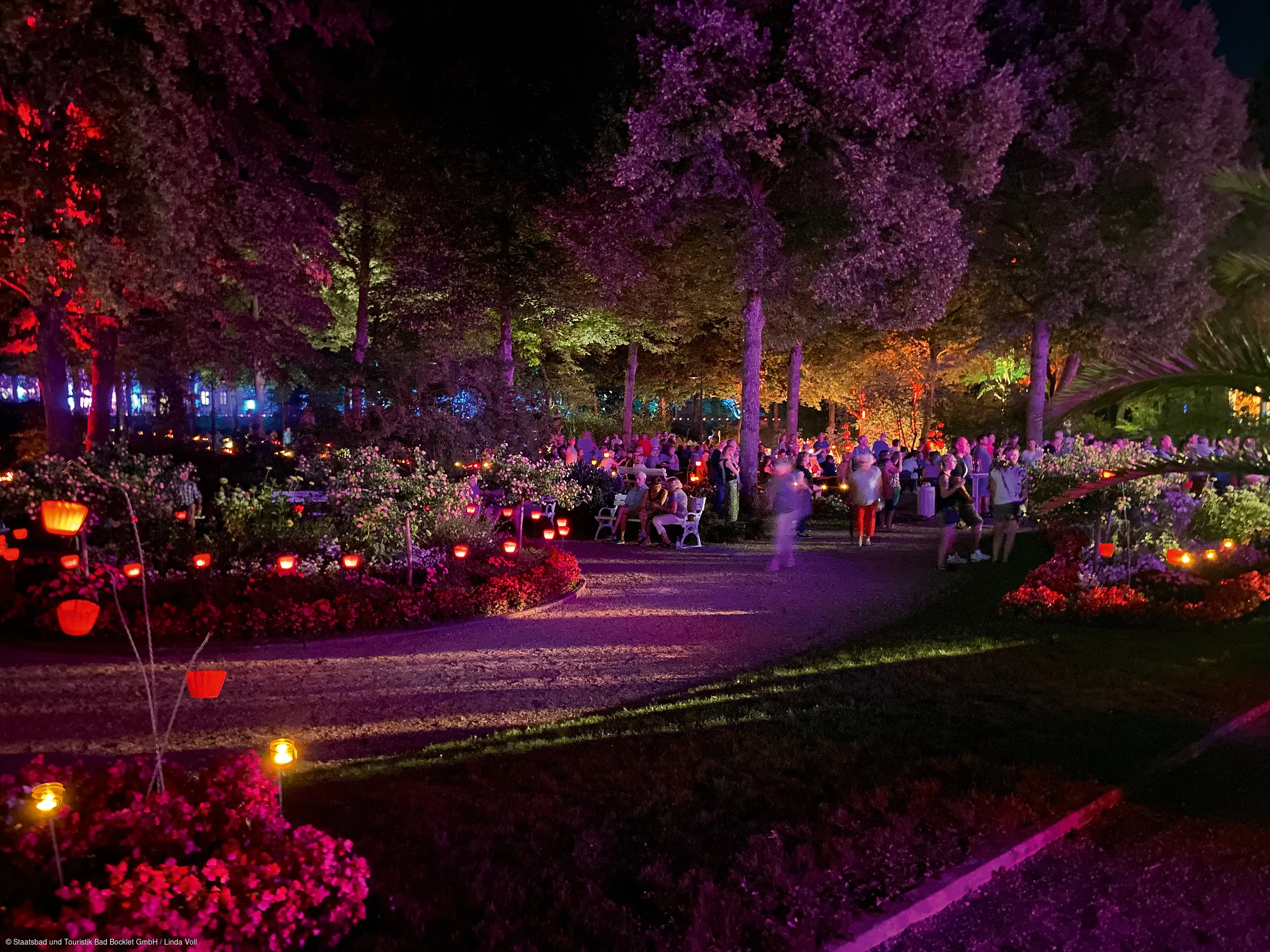 Parkweg bei Nacht mit bunten Lichtern, Blumenbeeten und vielen Menschen auf Bänken und stehend