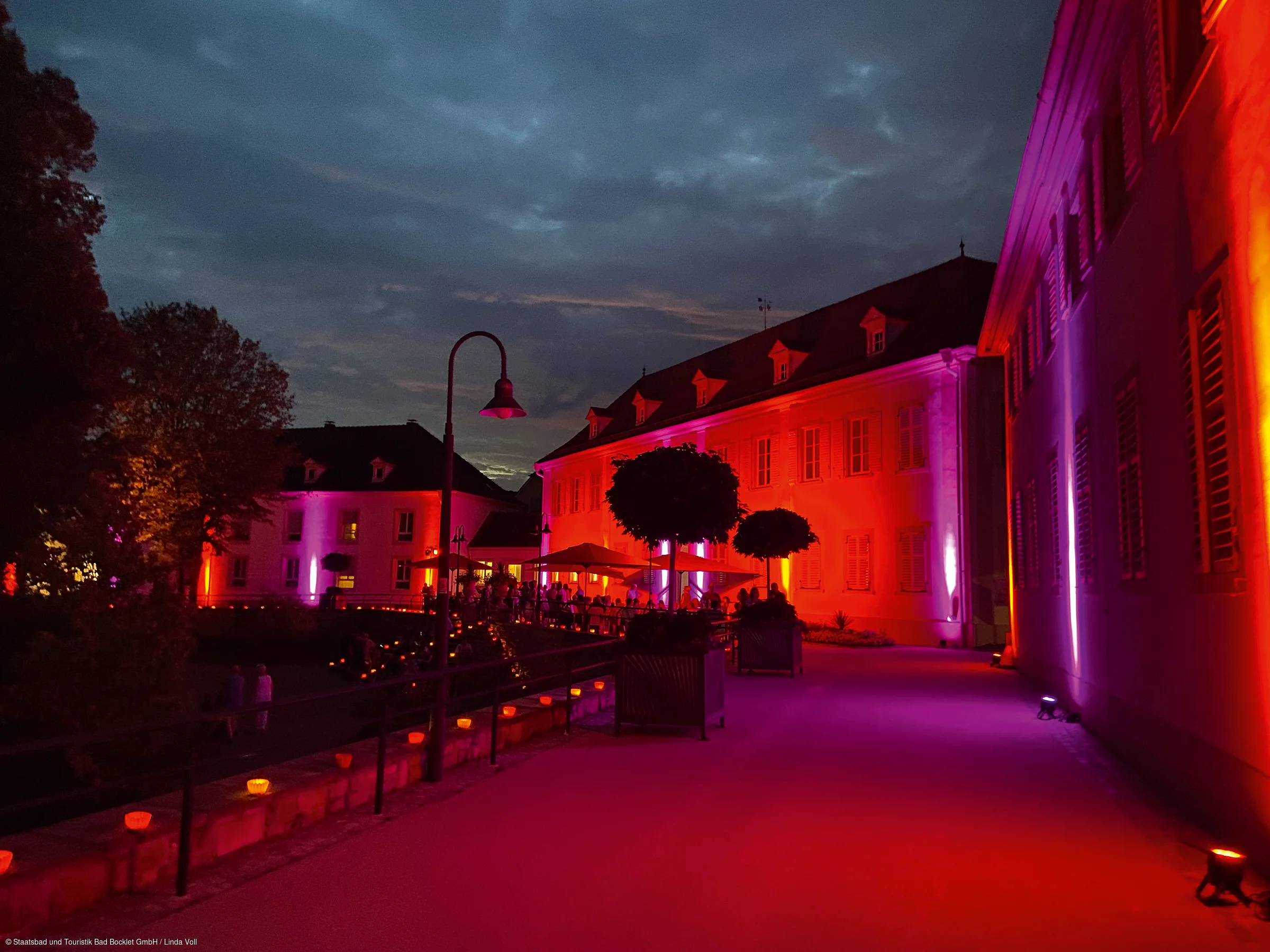Abendaufnahme eines beleuchteten Gebäudes mit roten und violetten Lichtern und Menschen auf der Terrasse.