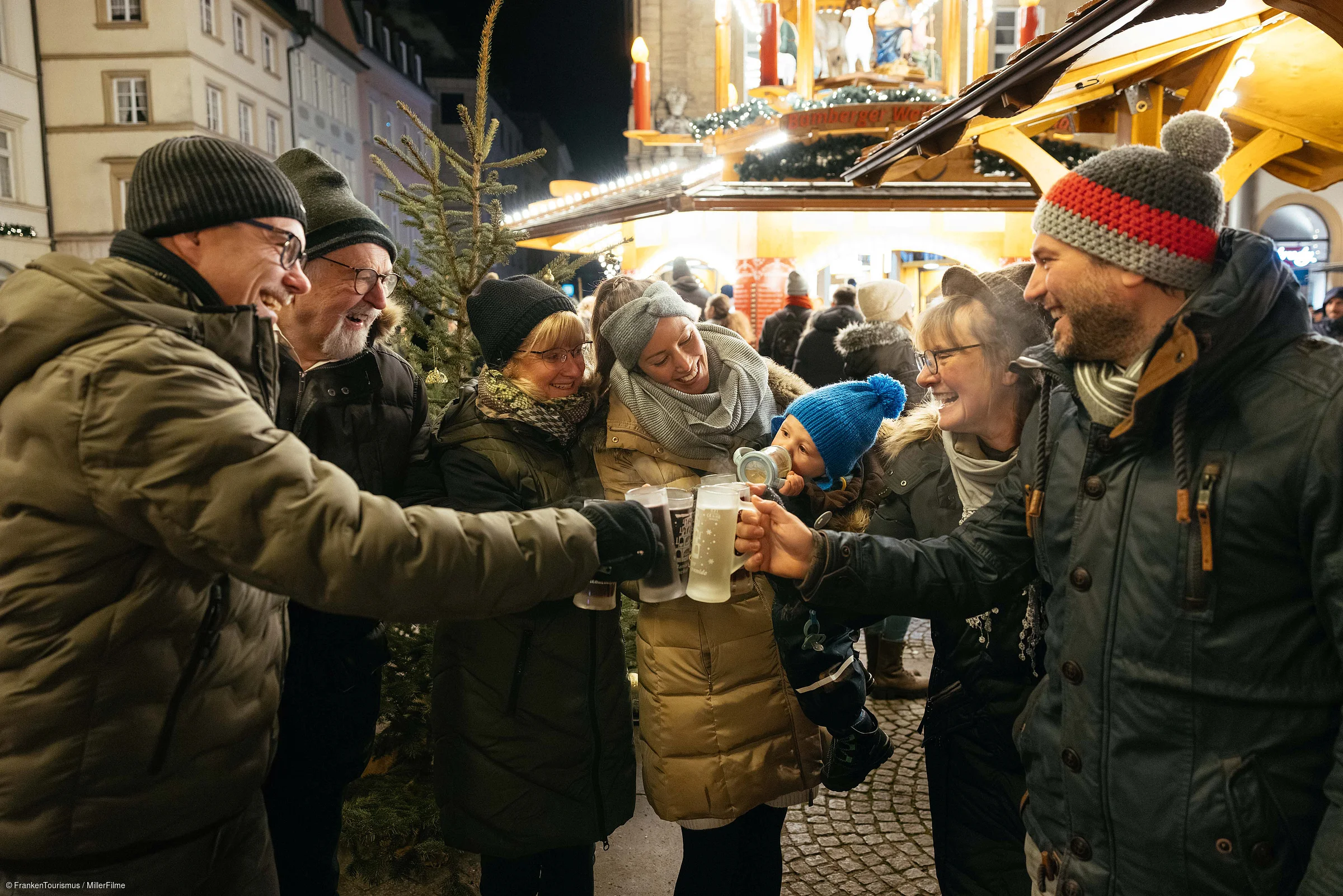 Mehrere Personen in Winterkleidung stoßen mit Getränkebecher auf einem Weihnachtsmarkt an.