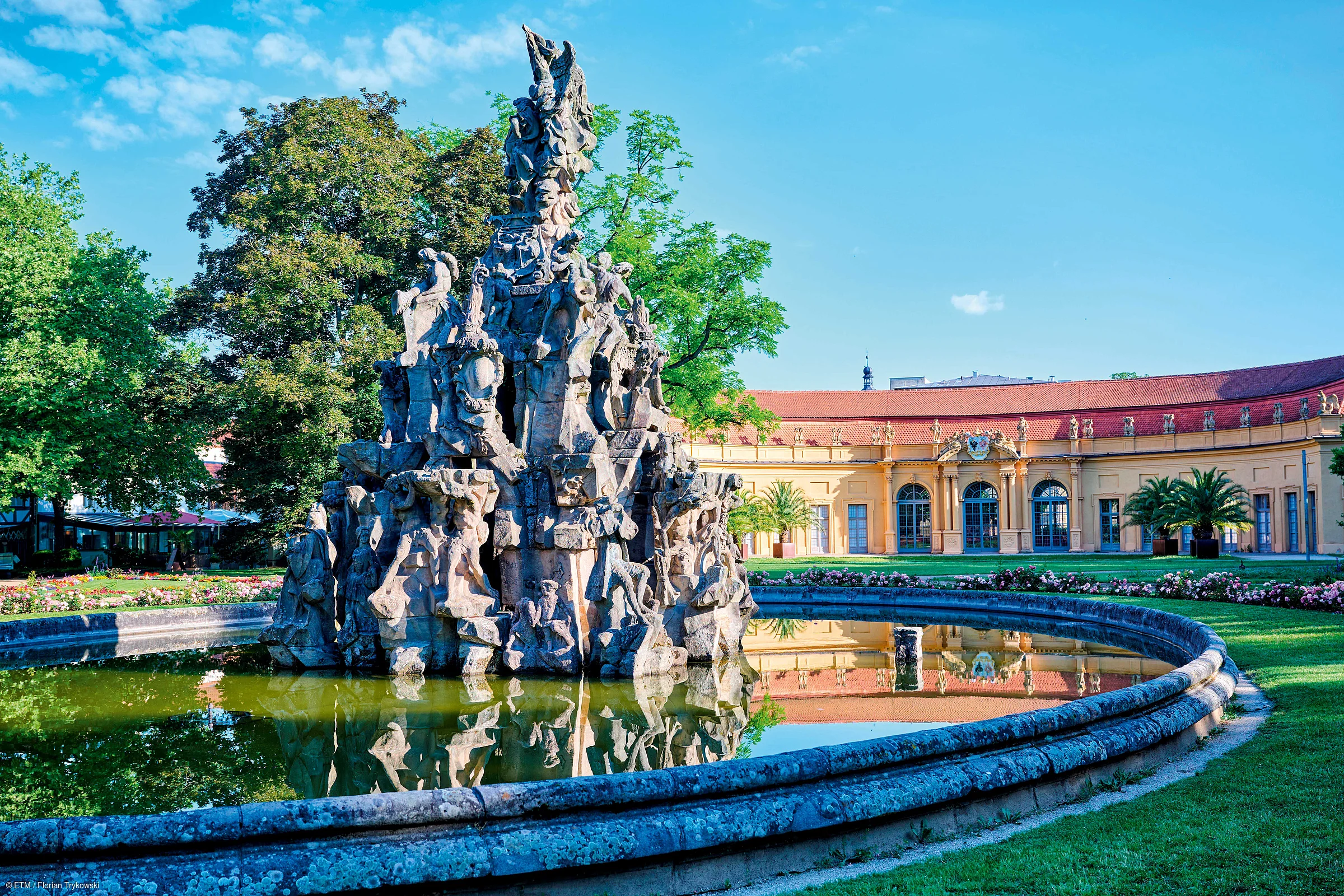 Steinerne Skulptur mit Figuren in rundem Wasserbecken vor gelbem Gebäude und Bäumen bei blauem Himmel.