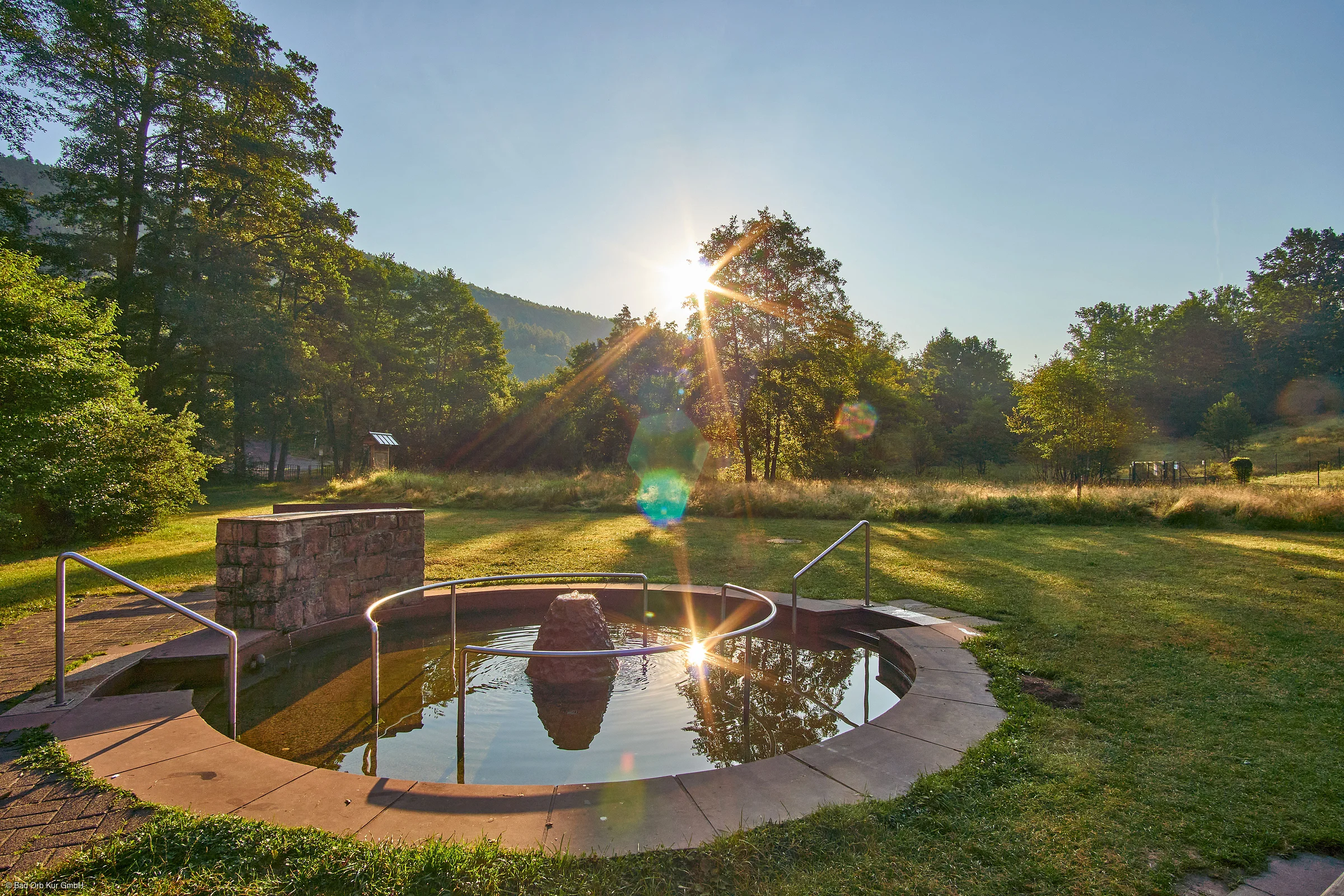 Kleiner runder Brunnen mit Wasser und Metallgeländer auf einer Wiese bei Sonnenuntergang im Wald.