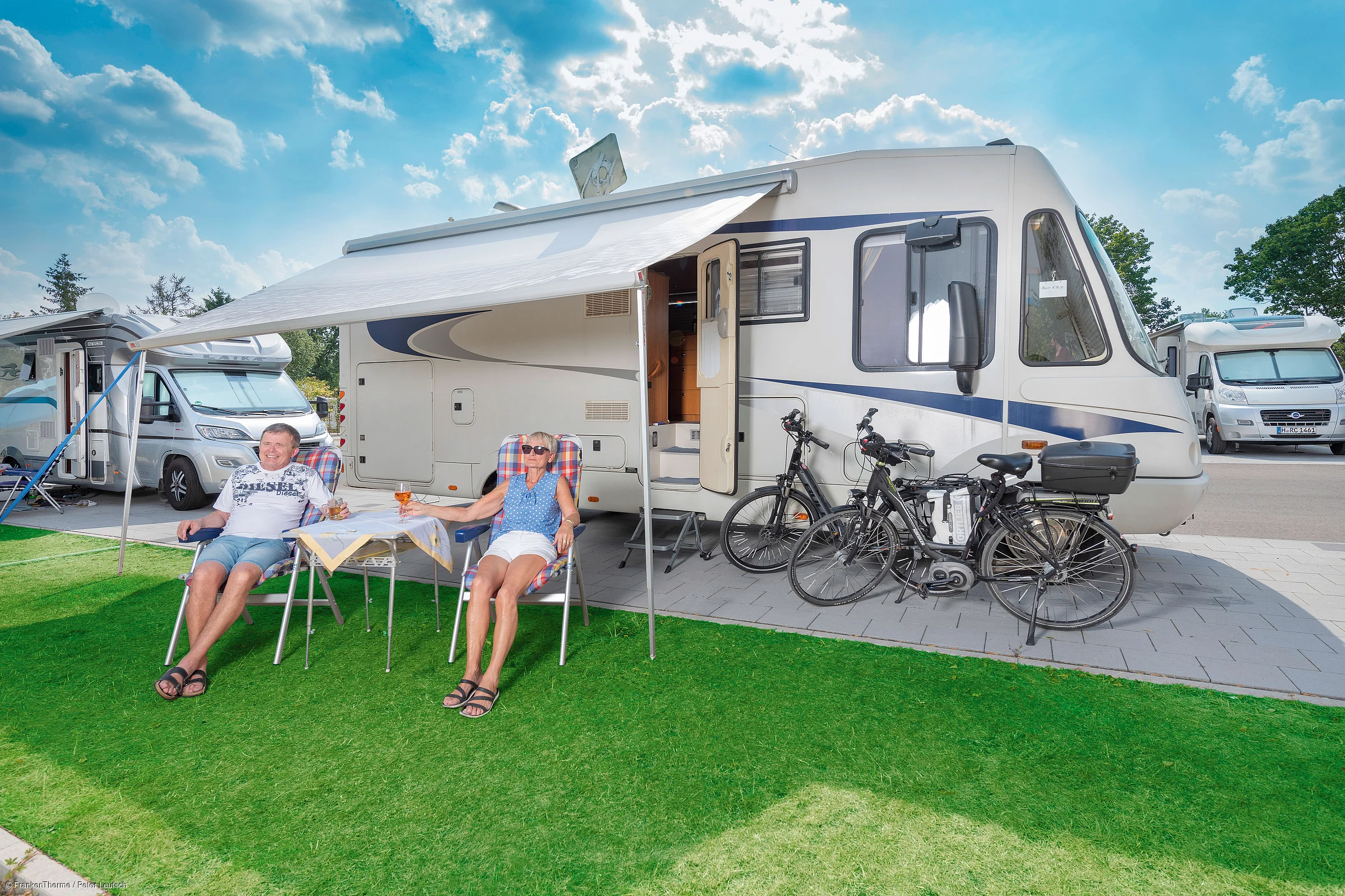Älteres Paar sitzt unter Markise vor Wohnmobil mit zwei Fahrrädern auf grünem Rasen bei sonnigem Himmel.