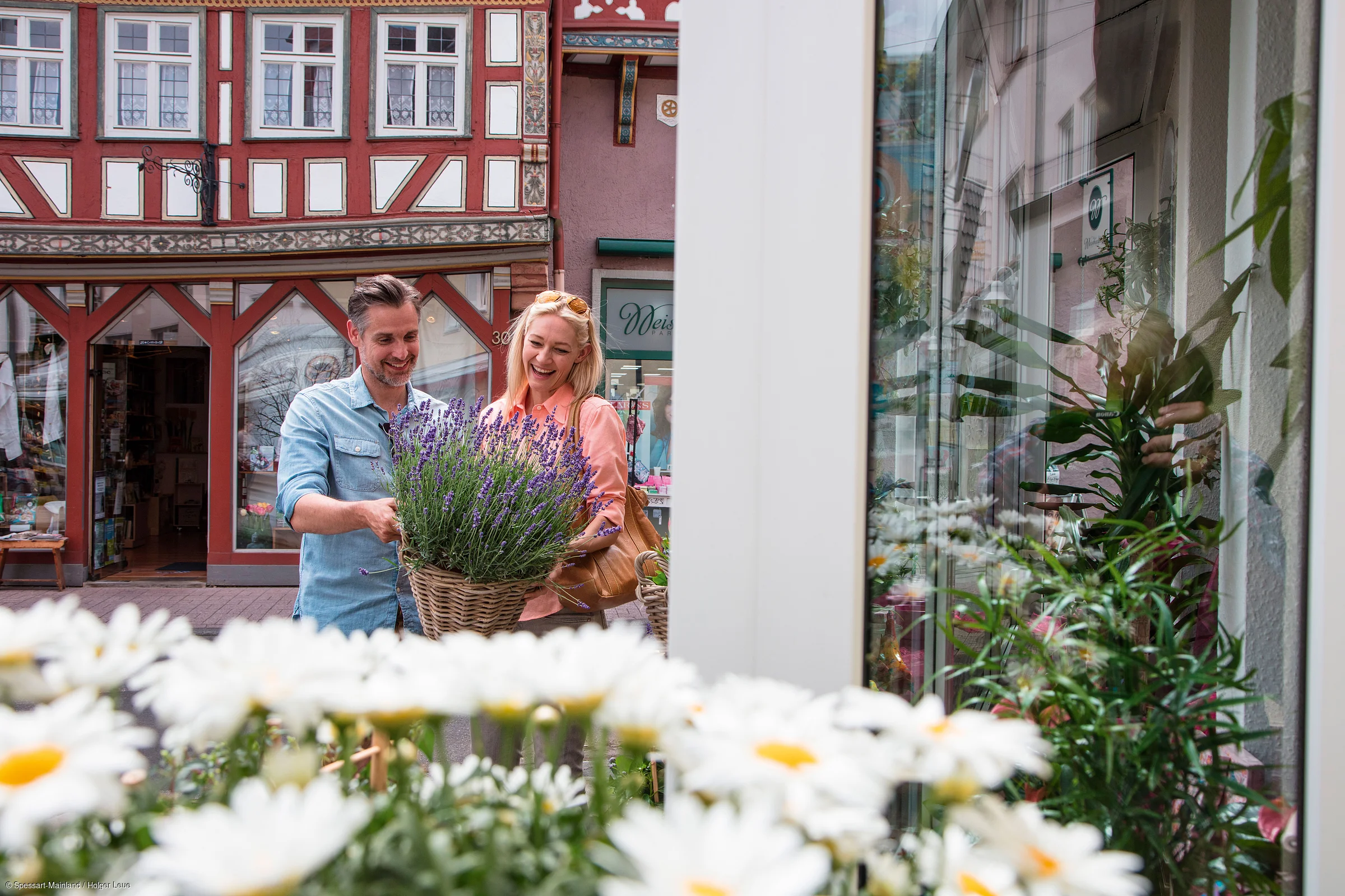 Paar betrachtet einen Korb mit Lavendel vor einem Fachwerkhaus und einem Schaufenster mit Pflanzen.