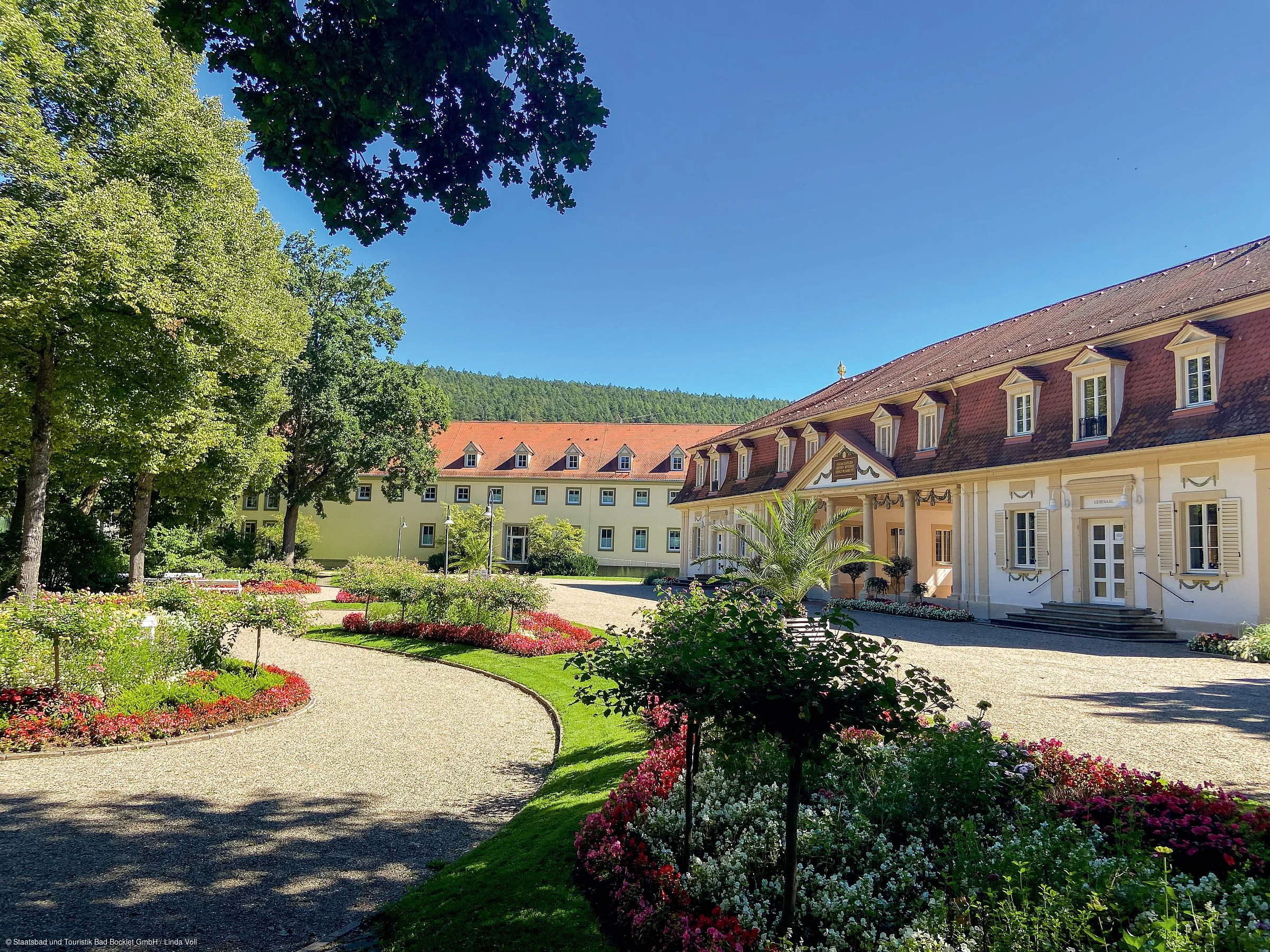 Garten mit Blumenbeeten, Bäumen und zwei Gebäuden mit roten Dächern unter blauem Himmel.