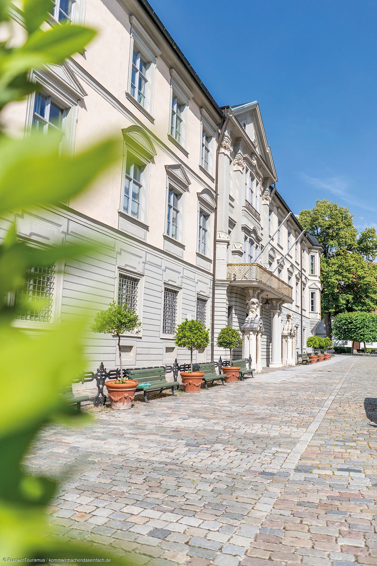 Fassade eines historischen Gebäudes mit Bänken und bepflanzten Töpfen auf gepflastertem Platz bei klarem Himmel.