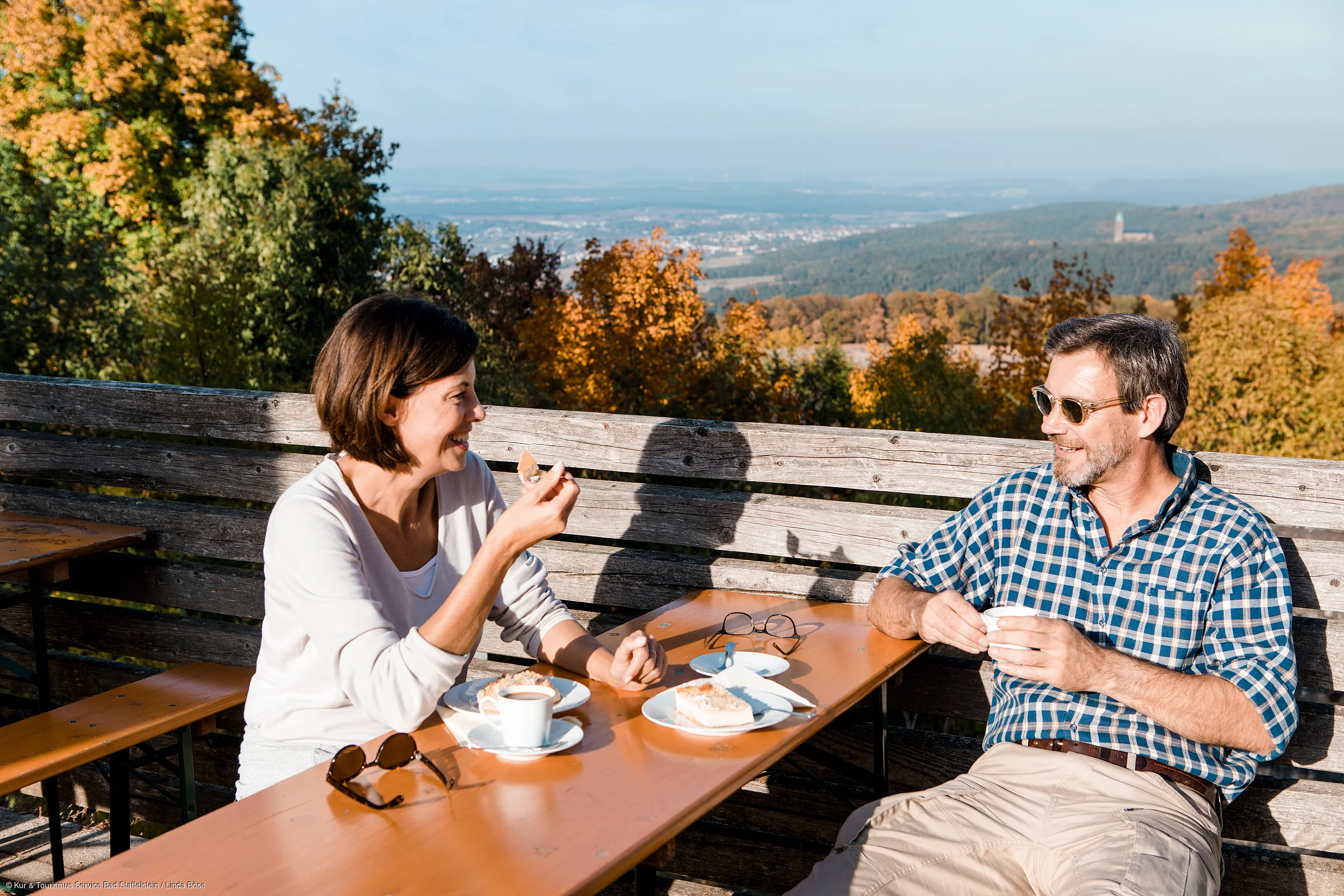 Zwei Personen sitzen draußen an einem Holztisch, essen Kuchen und trinken Kaffee, im Hintergrund Herbstwald.