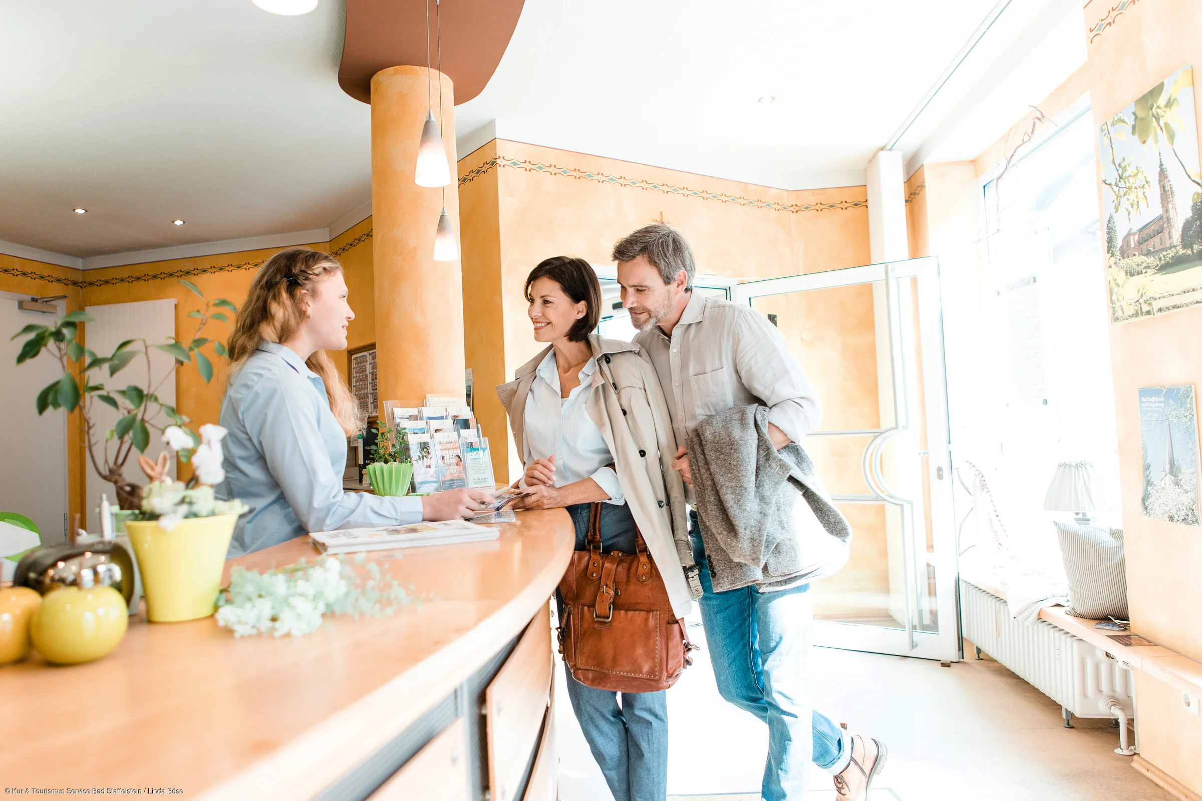 Paar steht an Rezeption und spricht mit Empfangsdame in hellem, freundlich eingerichteten Raum.