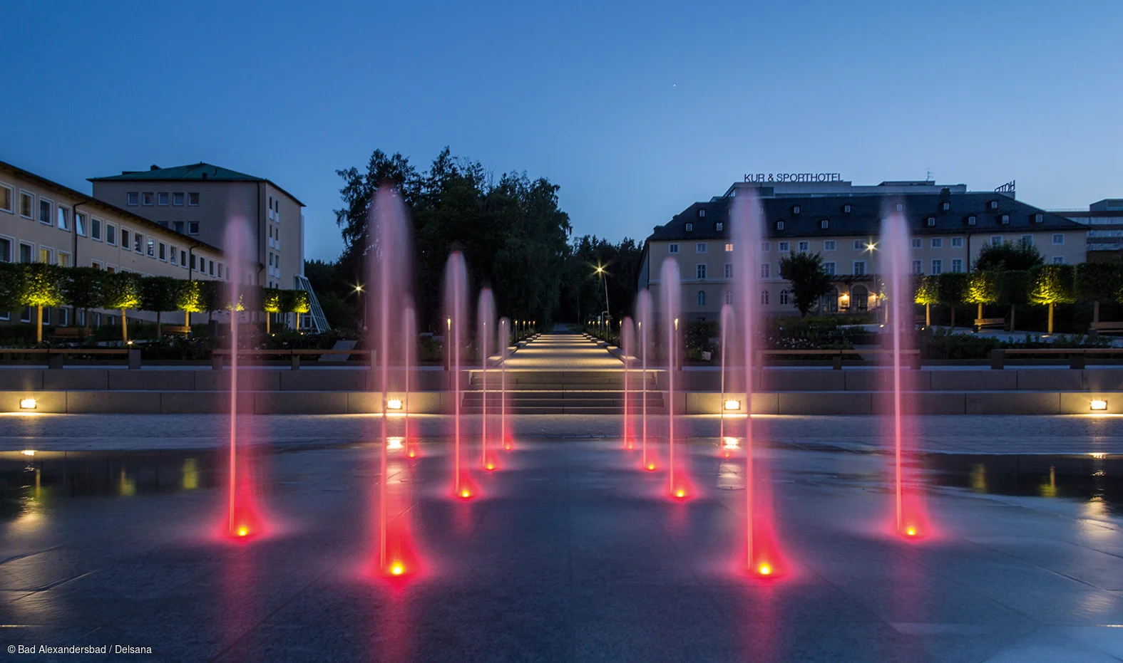 beleuchtete rote Wasserfontänen vor einem Gebäude bei Abenddämmerung mit blauem Himmel
