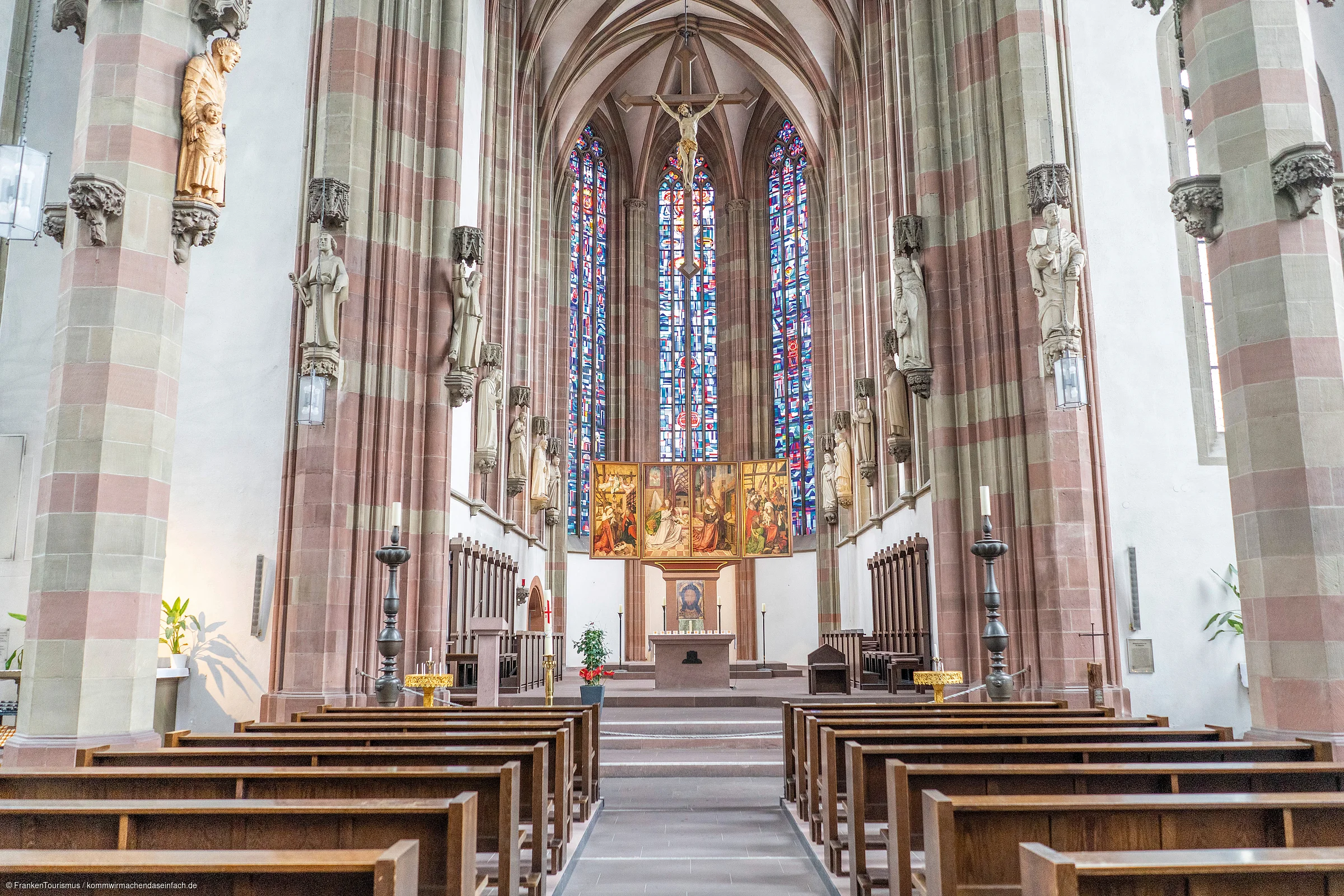 Innenraum einer Kirche mit Holzbänken, Altar, Kreuz und bunten Glasfenstern im Hintergrund