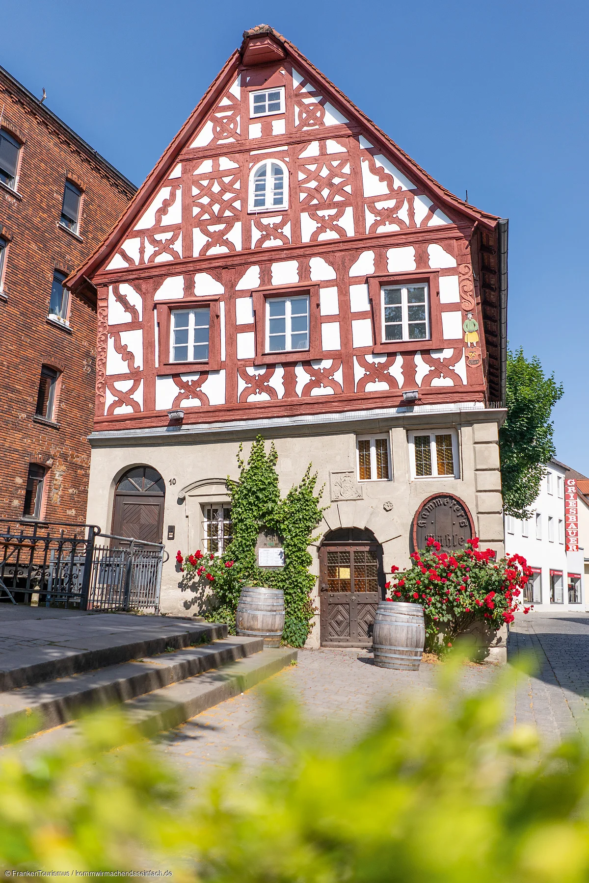 Fachwerkhaus mit roten Balken, weißen Wänden, zwei Holztüren, zwei Weinfässern und roten Blumen am Eingang.