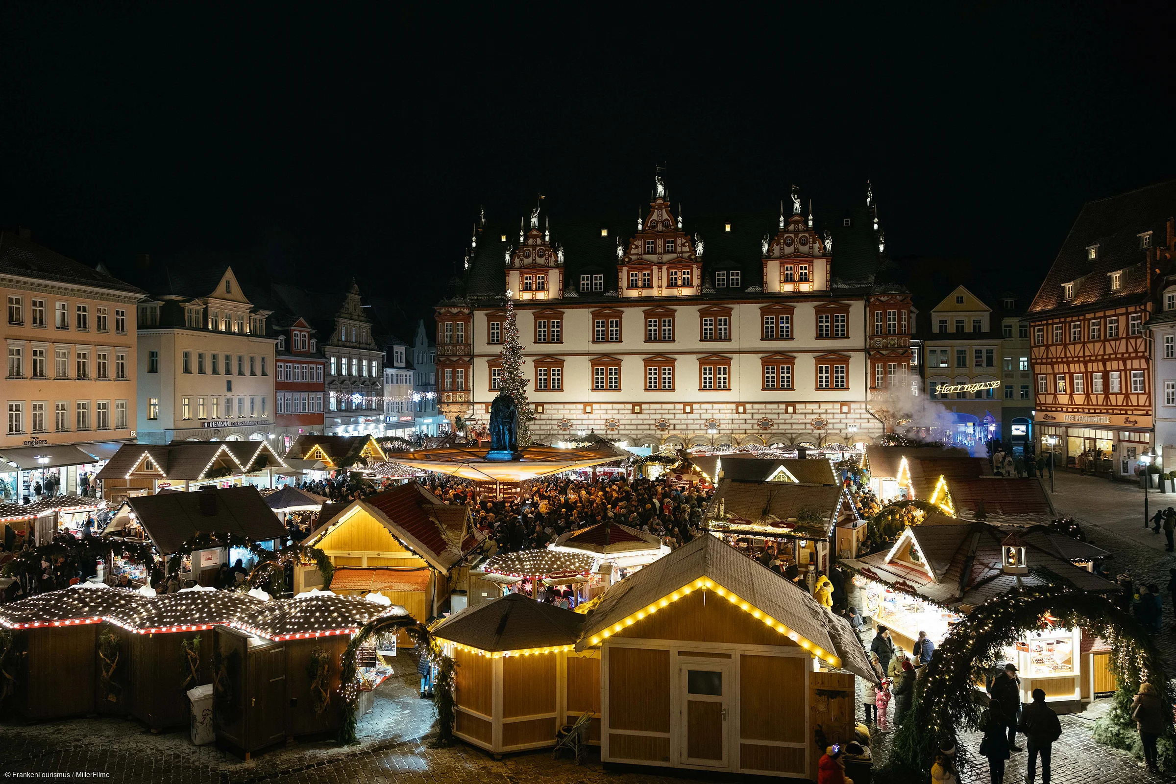 Weihnachtsmarkt mit beleuchteten Holzständen und vielen Besuchern vor historischem Rathaus bei Nacht.