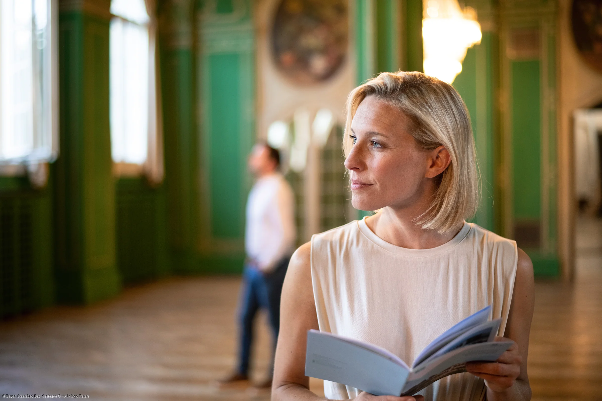 Frau mit kurzem blonden Haar hält ein geöffnetes Buch in einem großen Raum mit grünen Wänden.