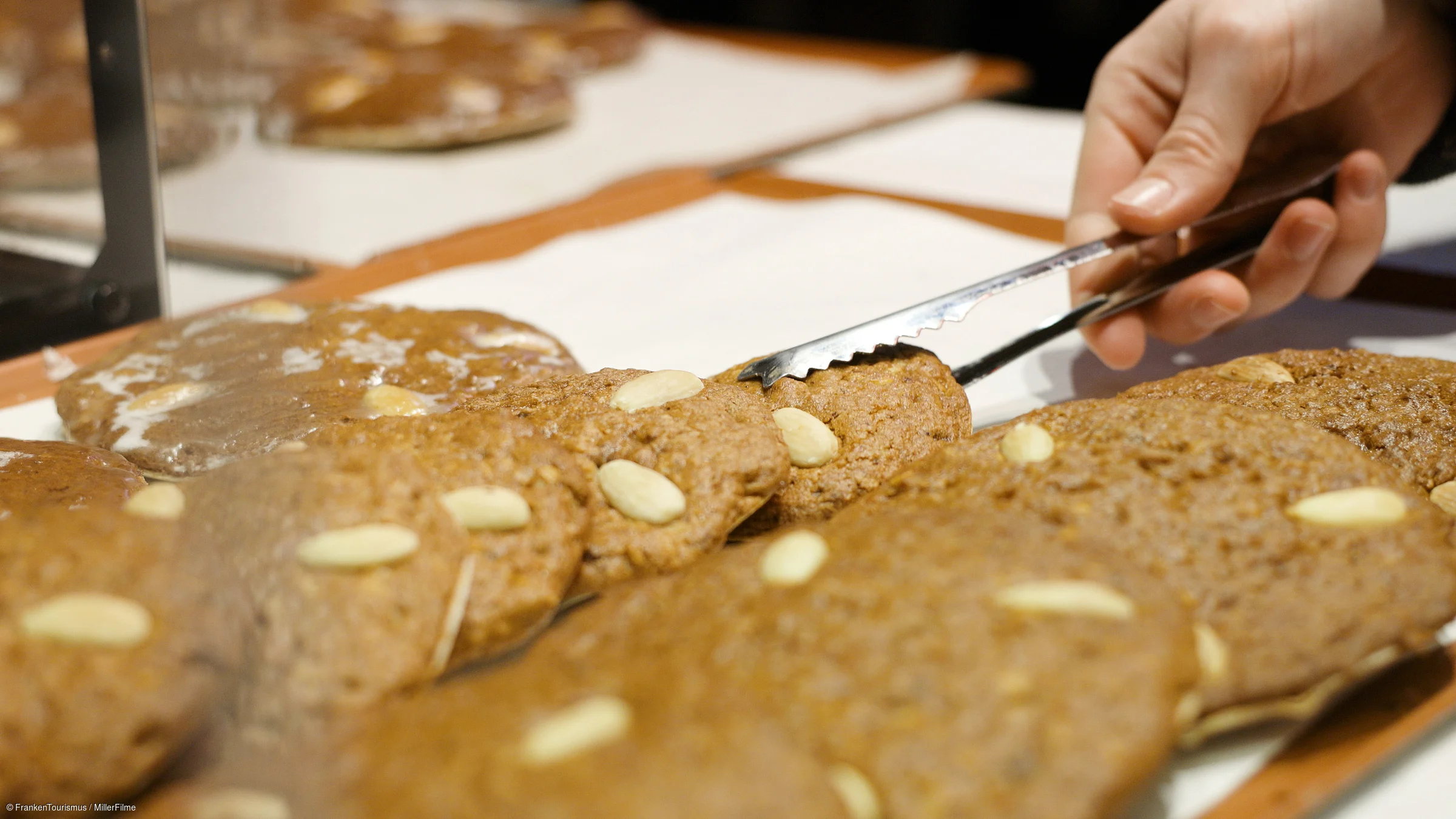 Hand mit Zange nimmt einen Lebkuchen mit Mandeln aus einer Auslage hinter Glas.