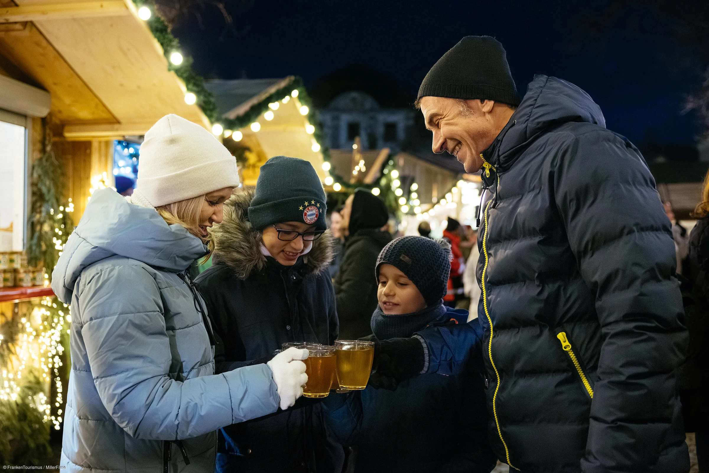 Familie mit Winterkleidung stößt mit Tassen bei Weihnachtsmarkt mit Lichterketten an.