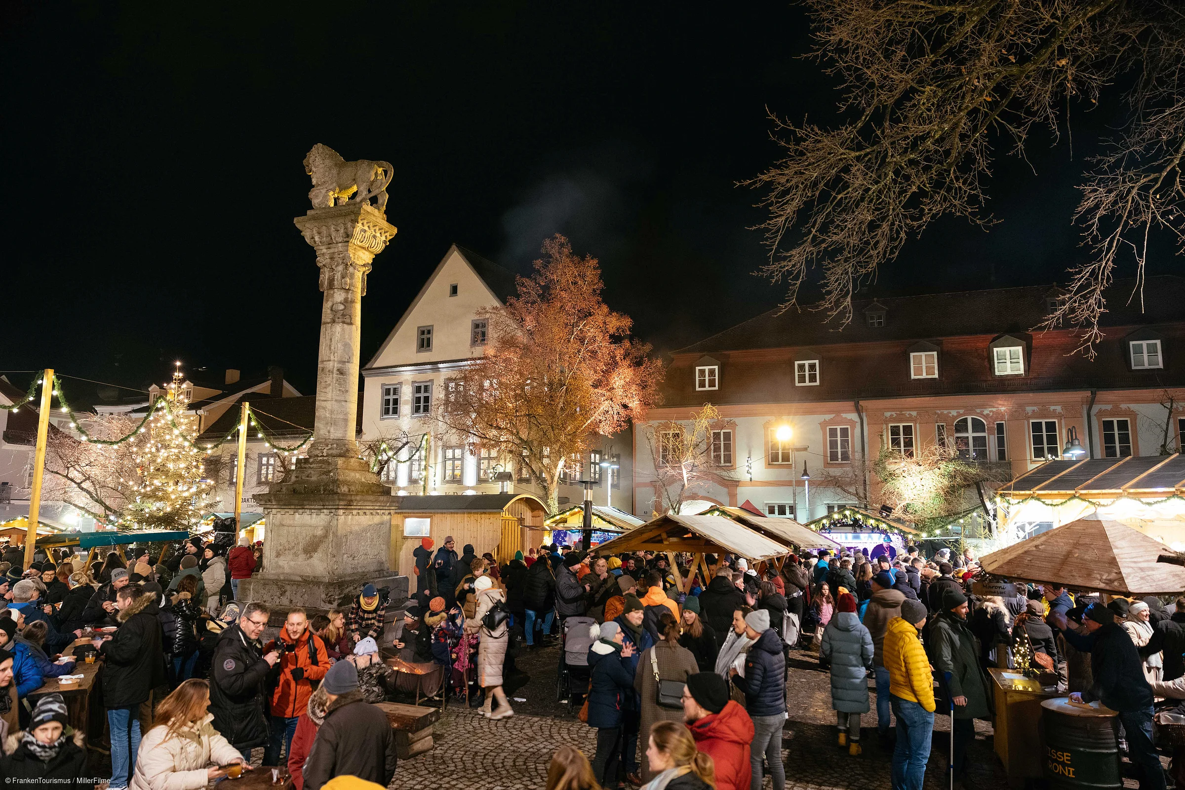 Weihnachtsmarkt mit vielen Menschen, beleuchteten Ständen und einer Säule bei Nacht.