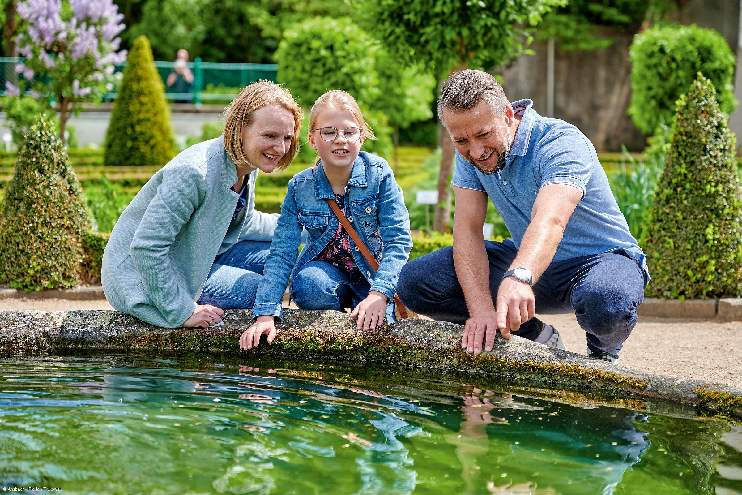 Drei Personen, zwei Erwachsene und ein Kind, sitzen am Rand eines Teichs und schauen ins Wasser in einem Garten.