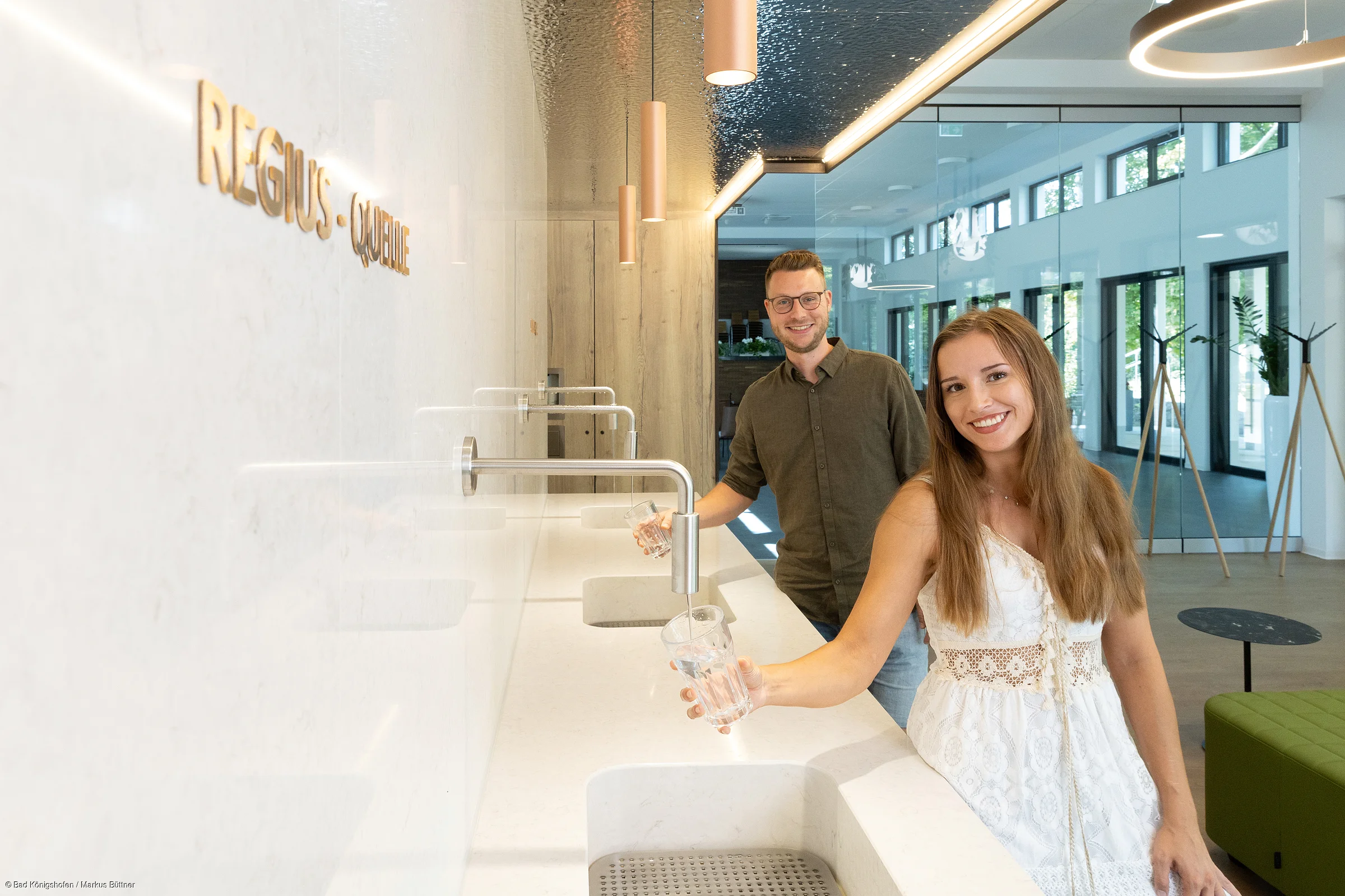 Zwei Personen füllen in modernem Innenraum an Wandwasserhähnen Gläser mit Wasser auf.