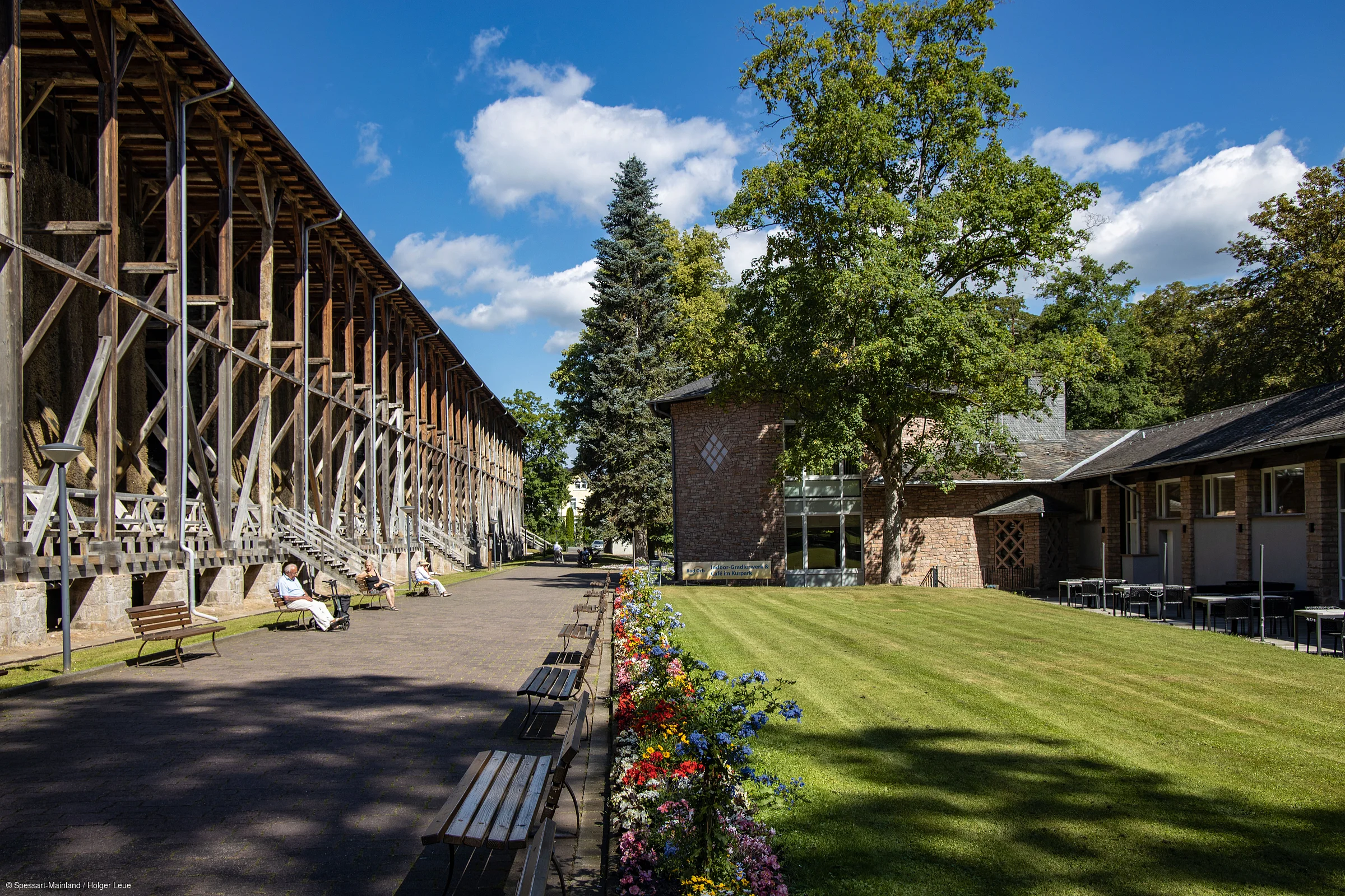 Außenbereich mit Holzkonstruktion, Sitzbänken, Blumenbeet und Gebäuden unter blauem Himmel mit Wolken.