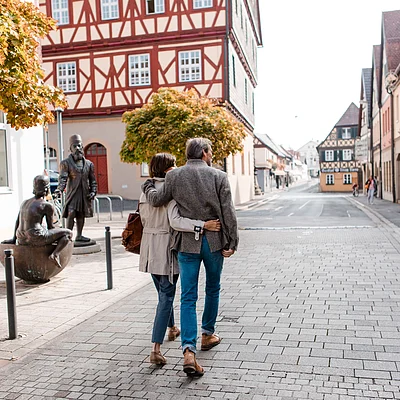 Paar geht Arm in Arm auf gepflasterter Straße in Altstadt mit Fachwerkhäusern und Skulpturen.