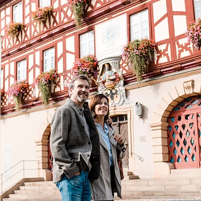Paar geht auf gepflastertem Platz vor Fachwerkhaus mit Blumen und roten Türen spazieren.