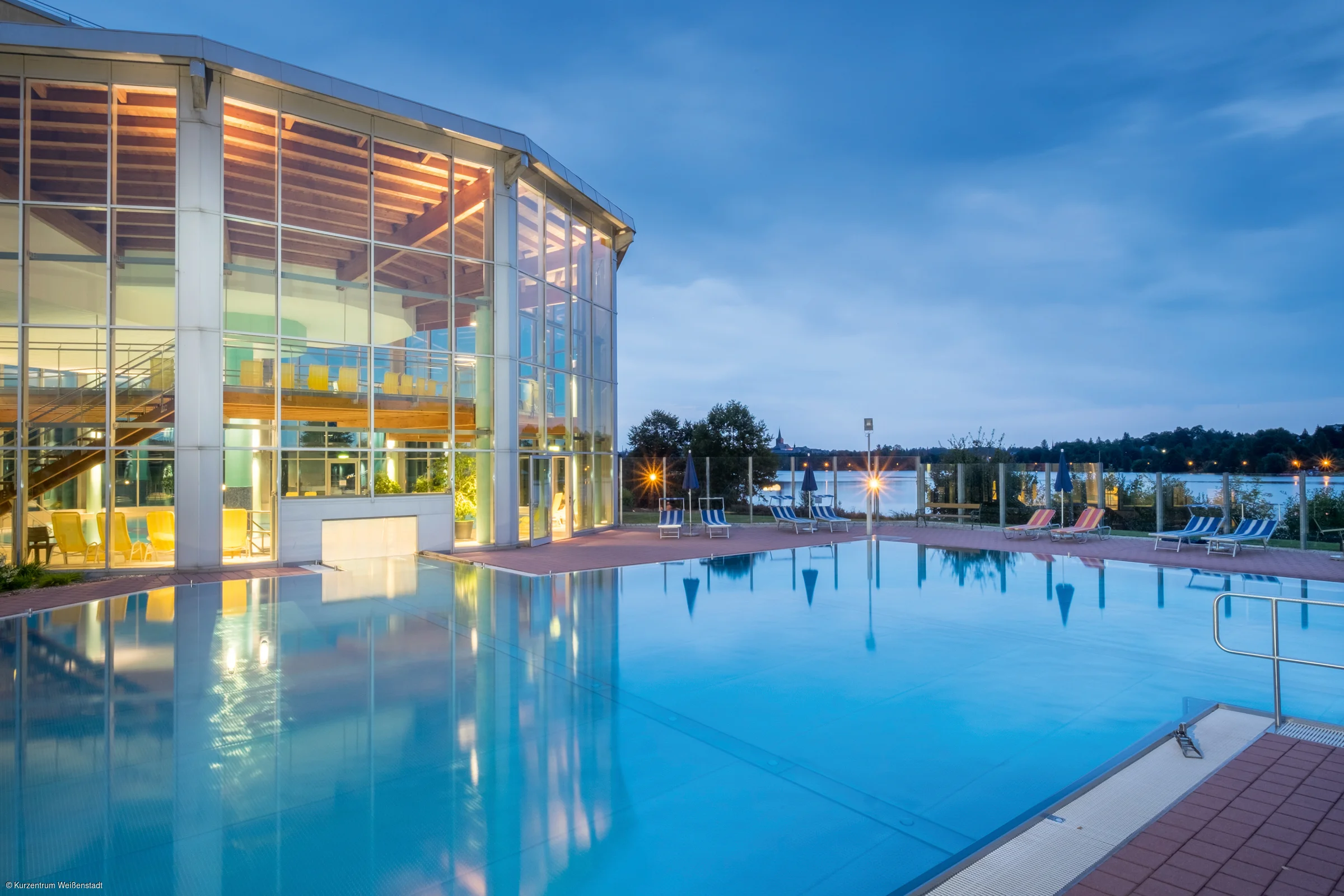 Außenpool bei Dämmerung neben einem großen Glasgebäude mit Innenbeleuchtung und Liegestühlen.