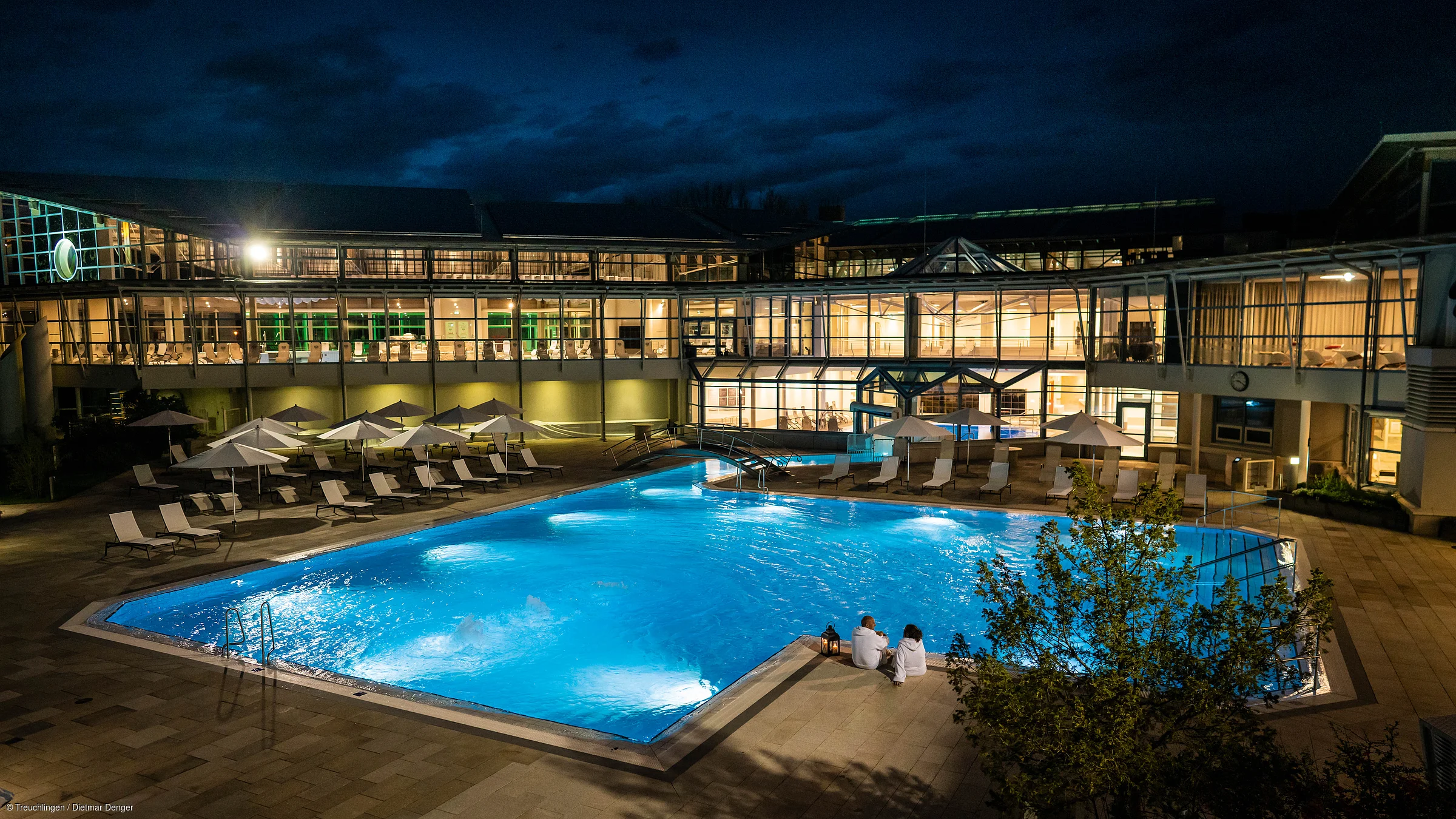 Beleuchtetes Schwimmbecken mit Liegestühlen und Sonnenschirmen vor einem modernen Gebäude bei Nacht.