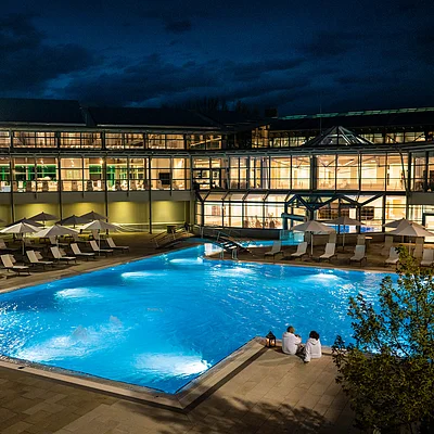 Beleuchtetes Schwimmbecken mit Liegestühlen und Sonnenschirmen vor einem modernen Gebäude bei Nacht.