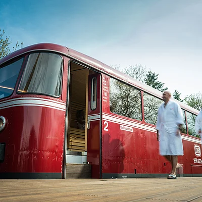 Roter Zug als Sauna mit offener Tür auf Holzbahnsteig, zwei Personen in weißen Bademänteln gehen vorbei.