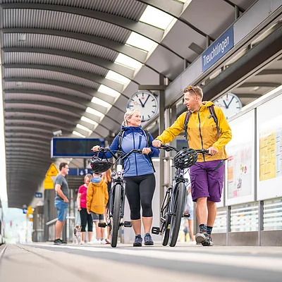 Zwei Personen mit Fahrrädern gehen auf einem Bahnsteig am Bahnhof Treuchtlingen, Zug links im Bild.