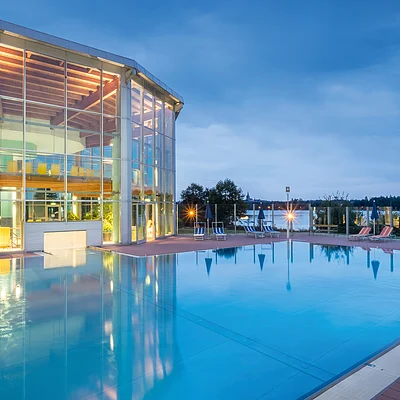 Außenpool bei Dämmerung neben einem großen Glasgebäude mit Innenbeleuchtung und Liegestühlen.