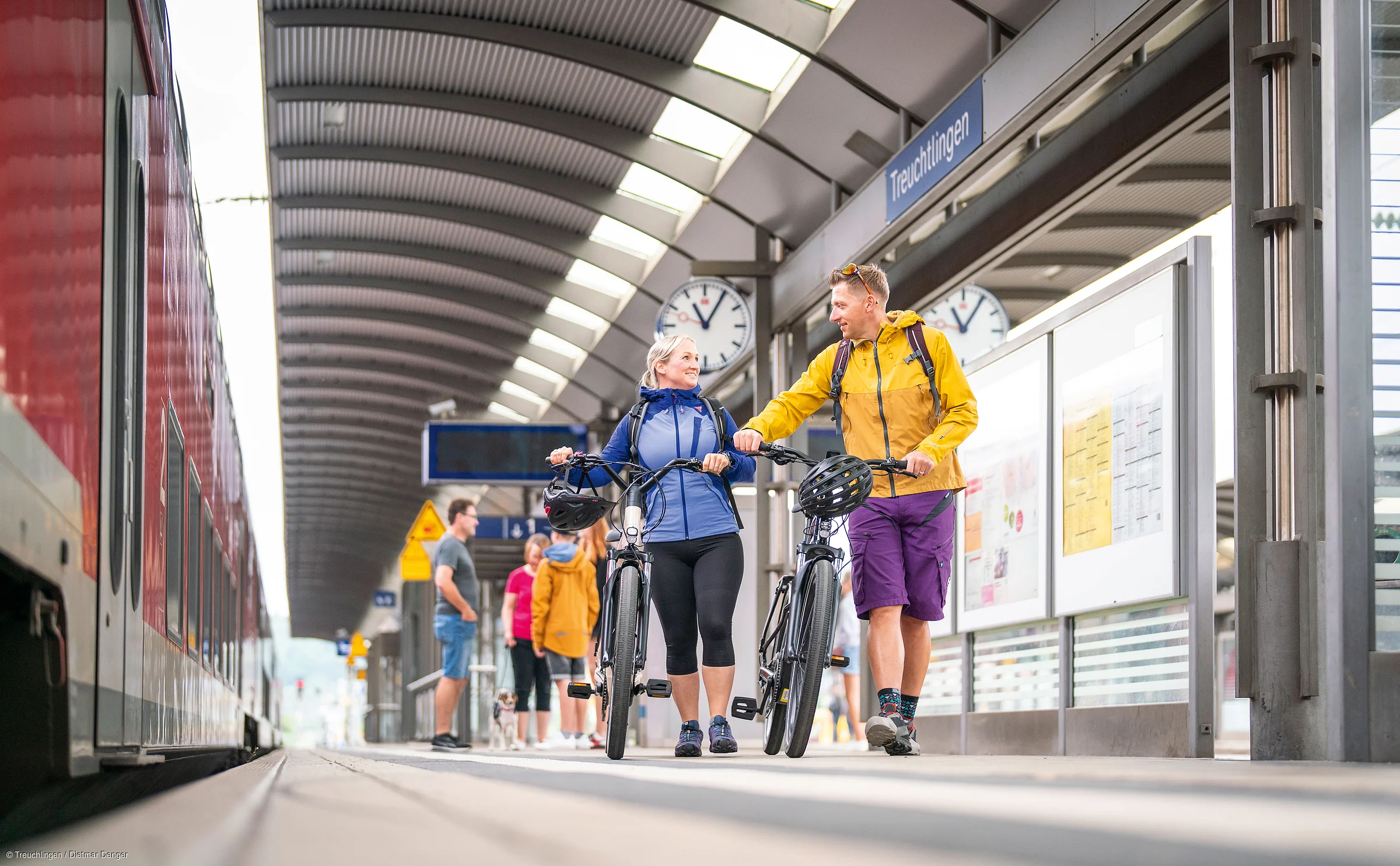 Zwei Personen mit Fahrrädern gehen auf einem Bahnsteig am Bahnhof Treuchtlingen, Zug links im Bild.
