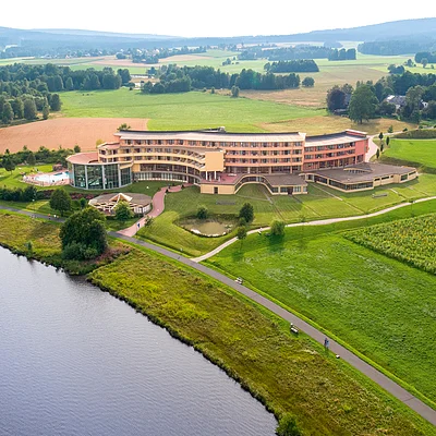 Luftaufnahme eines großen Gebäudes an einem Seeufer, umgeben von Wiesen, Feldern und Bäumen.